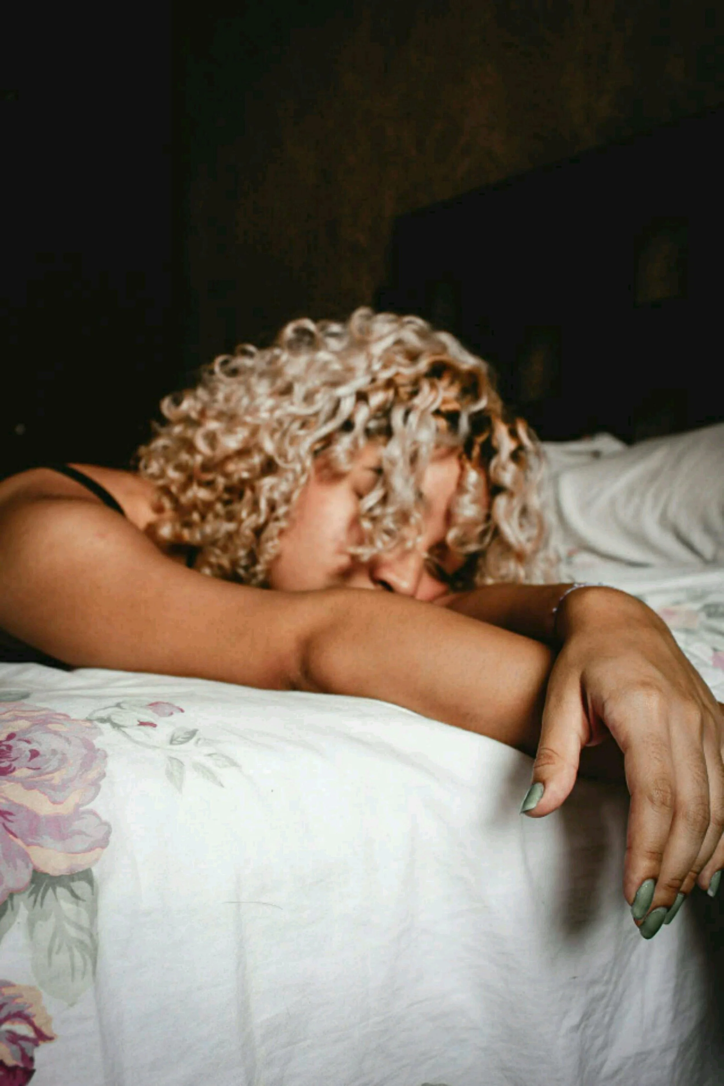 A woman with curly blonde hair sleeping on a bed with floral sheets, arm stretched out and head resting on her arm.