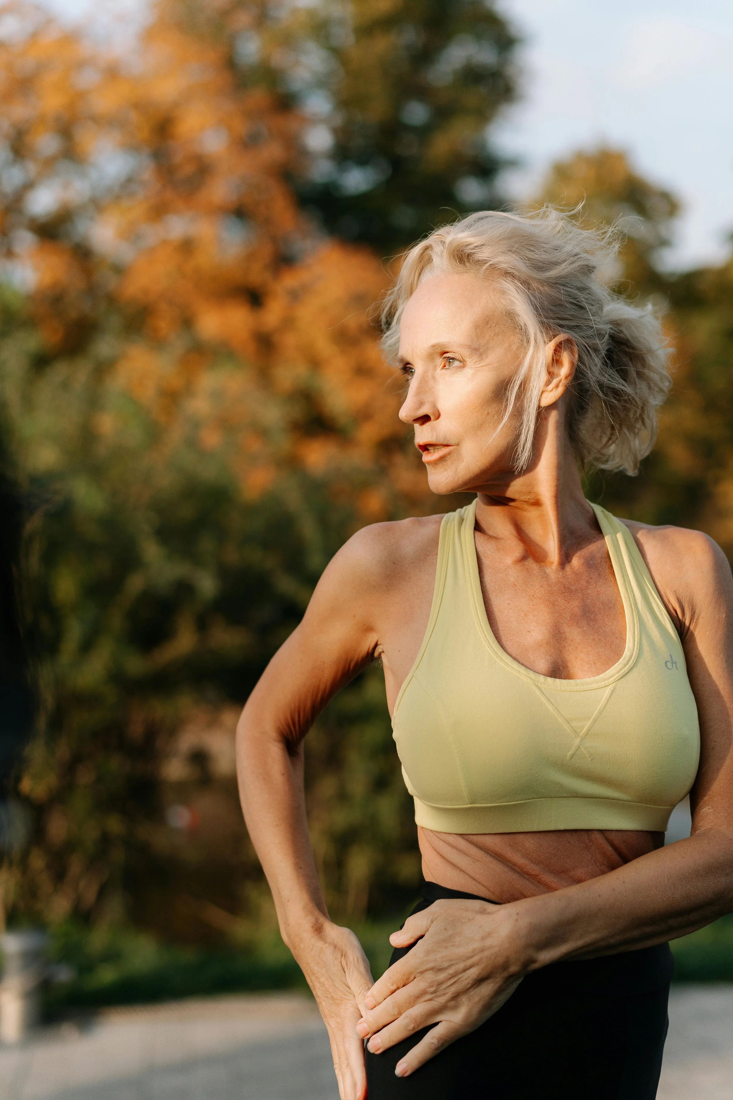 An older woman with gray hair wearing a yellow sports bra and black workout pants, standing outdoors with trees in the background during sunset.
