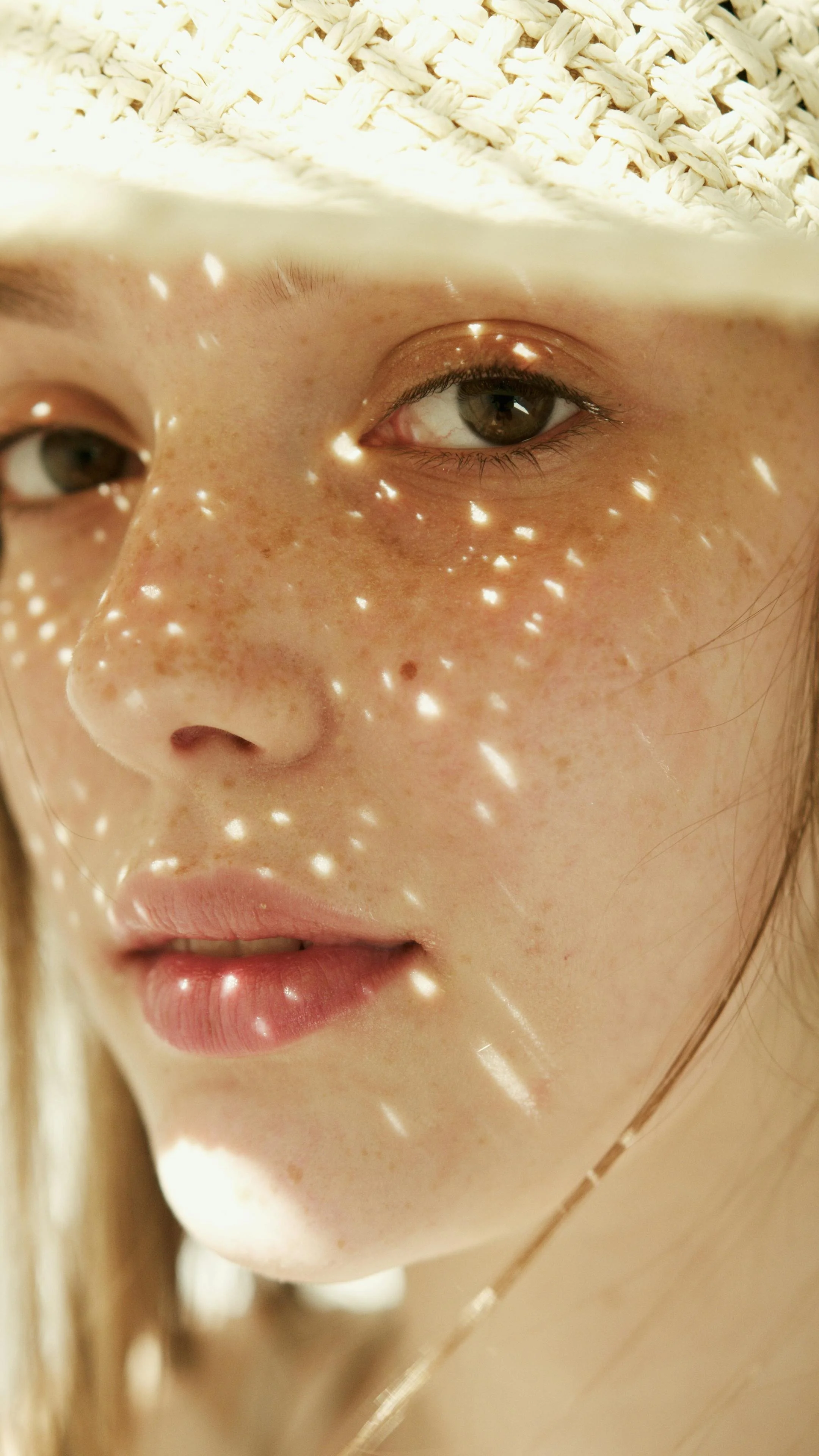 Close-up of a woman's face with freckles, partially covered by a woven hat, with sunlight creating dotted shadows on her skin.