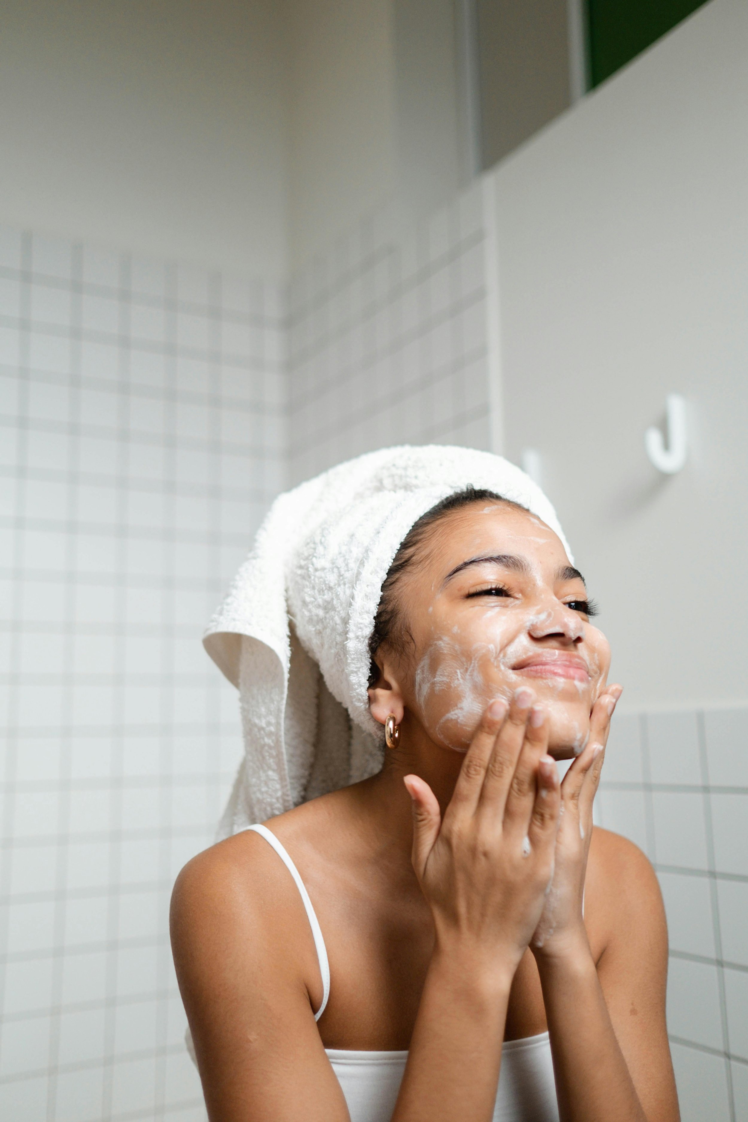 A young woman with a towel wrapped around her hair, smiling, as she applies face wash in a bathroom.