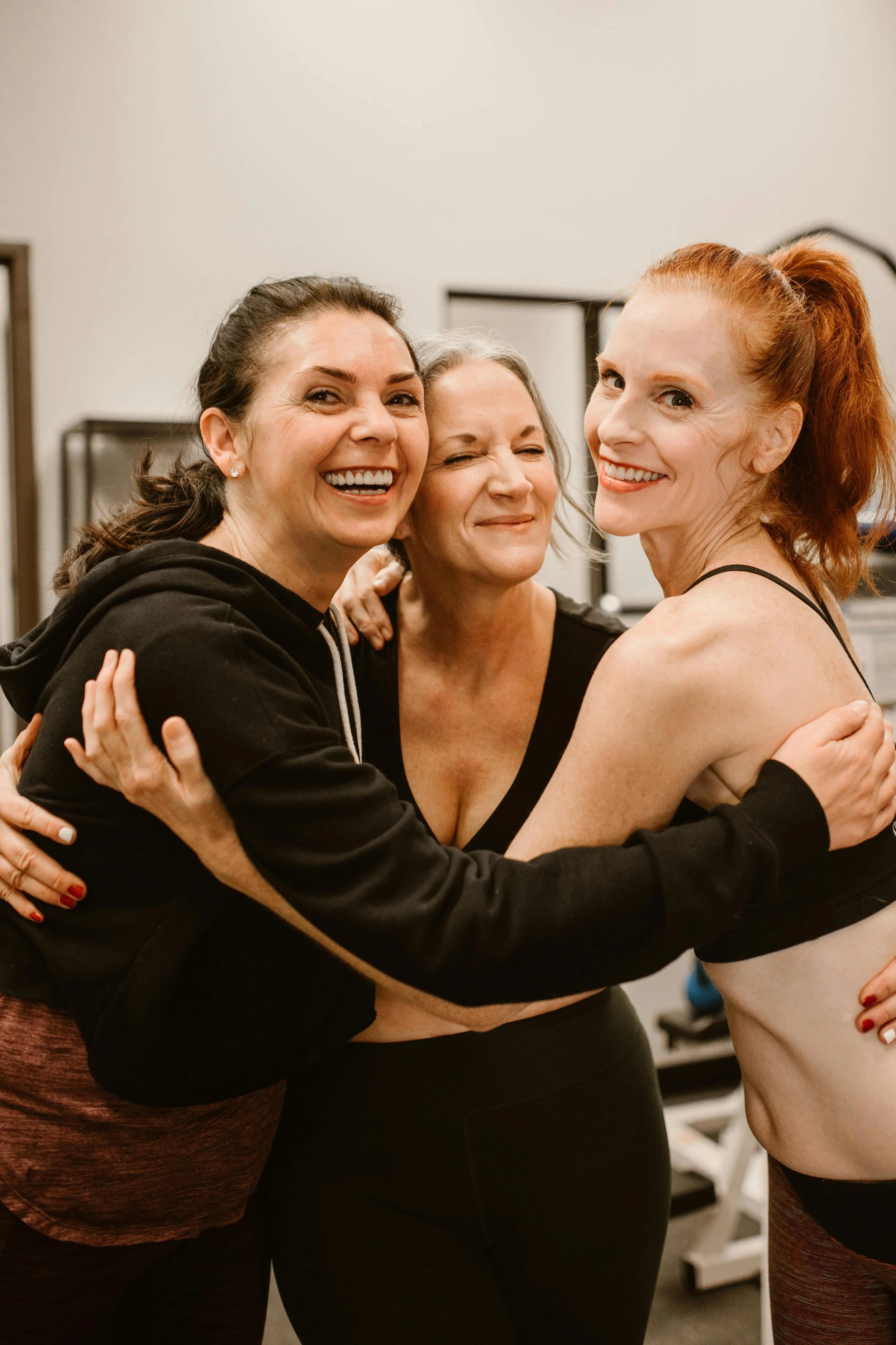 Three women hugging and smiling at the gym, one wearing a black hoodie, another with gray hair, and the third with red hair in a ponytail, wearing workout clothes.