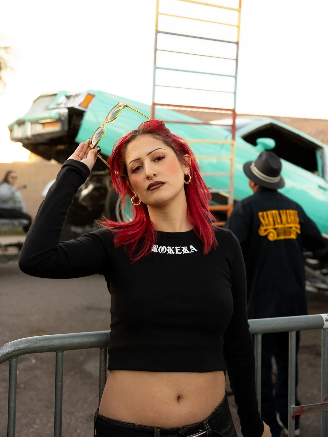 A woman with red hair and dark makeup poses confidently in front of a crashed turquoise airplane at an outdoor event, holding sunglasses in her hand and wearing a black cropped top with white text.