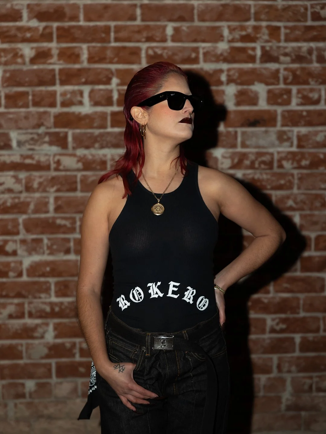 A woman with red hair, wearing black sunglasses, a black tank top with 'ROKER' written on it, dark jeans, gold jewelry, in front of a brick wall.