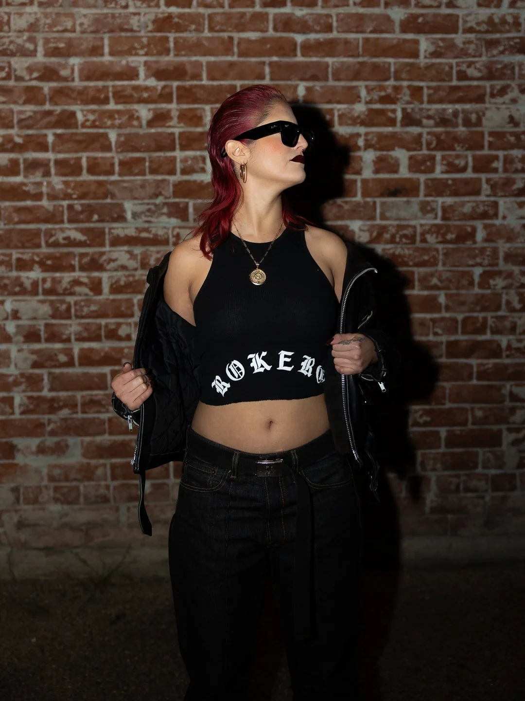 Woman with red hair wearing sunglasses, gold jewelry, black crop top with white "ROKEBO" text, black pants, and a black jacket draped off her shoulders, standing against a brick wall.