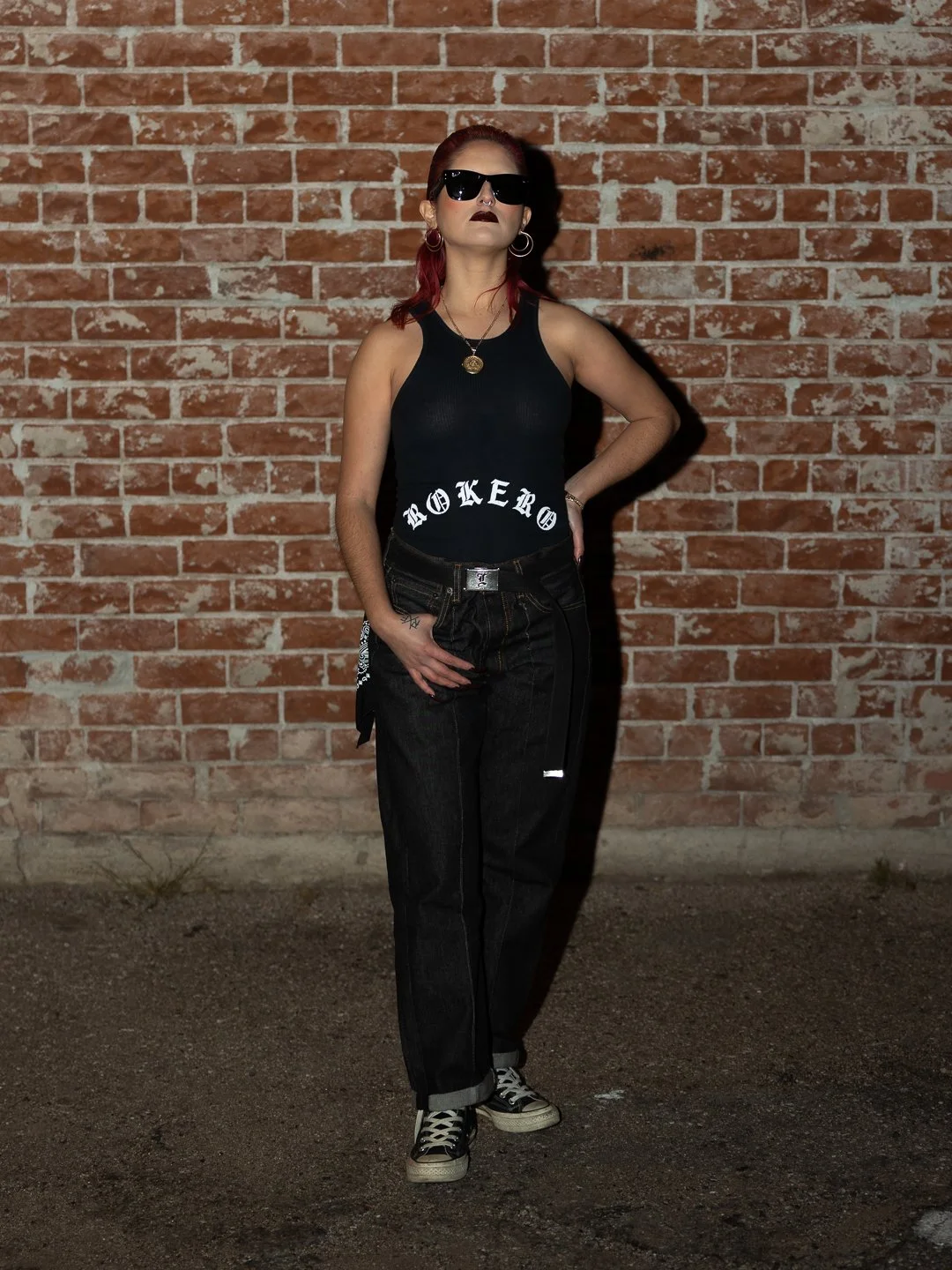 A woman with red hair and dark makeup stands against a brick wall, wearing sunglasses, a black tank top with 'BROKER' written on it, black jeans, and sneakers.