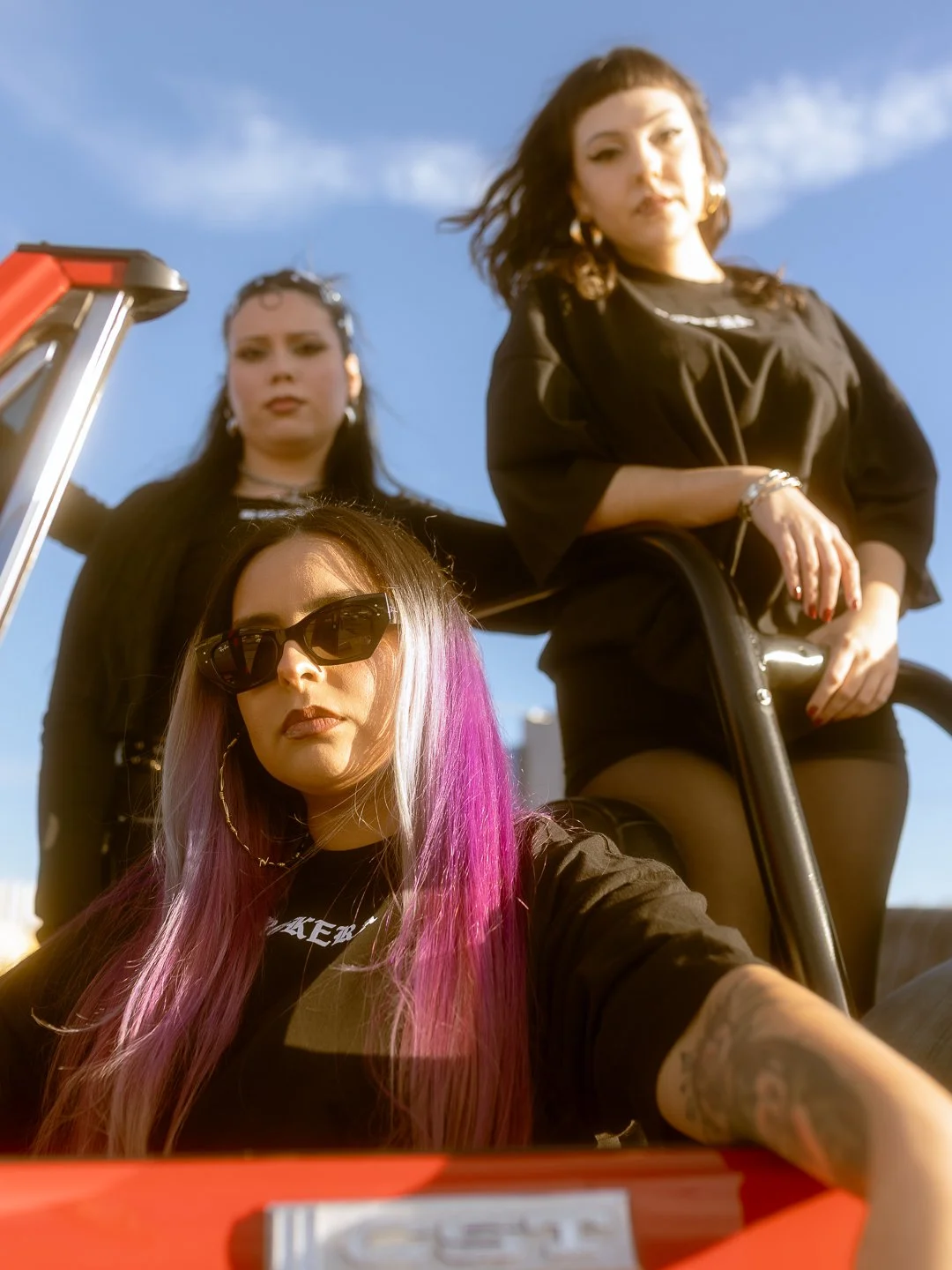 Three women posing outdoors against a blue sky. One is sitting in a vehicle with pink and blonde hair, wearing sunglasses. The other two are standing behind, dressed in black, with one having dark wavy hair and the other with dark straight hair.