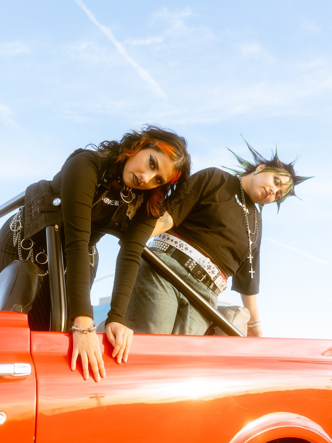 Two young people with punk style hair and accessories leaning over the door of a red convertible car against a blue sky.