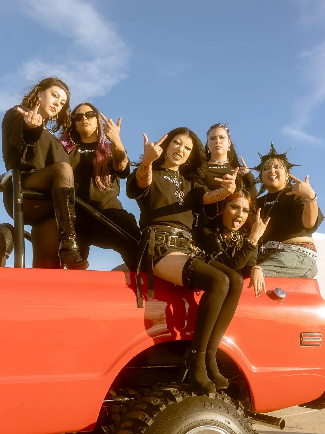 Group of six young women with alternative fashion posing on top of a red vehicle against a blue sky, making rebellious hand gestures and facial expressions.