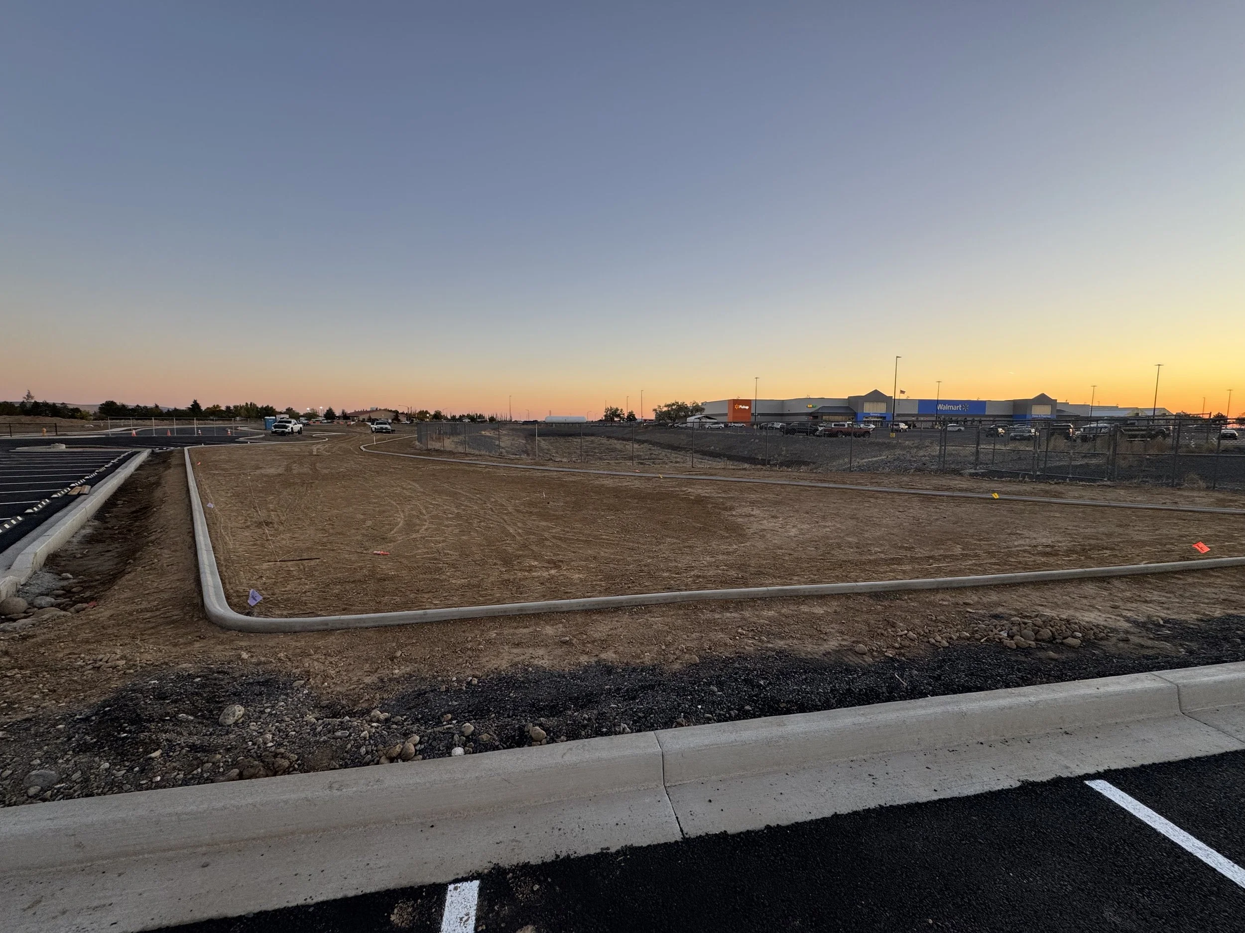 Empty parking lot under construction with dirt ground, fencing, and a Walmart store in the background at sunset.
