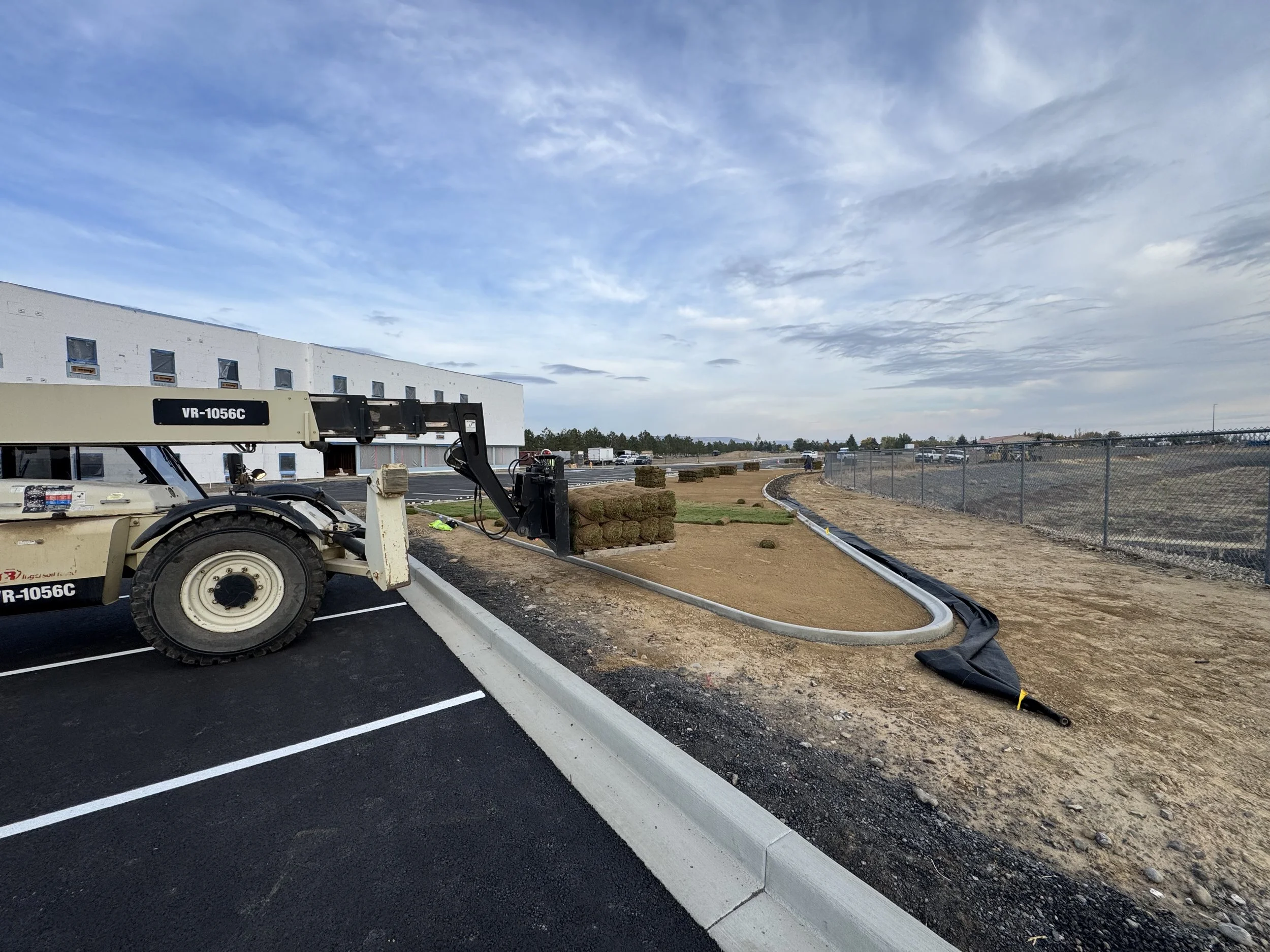 Construction site with a vehicle and a curved concrete edge, black fabric liner, and an open field with industrial buildings and a fence in the background.