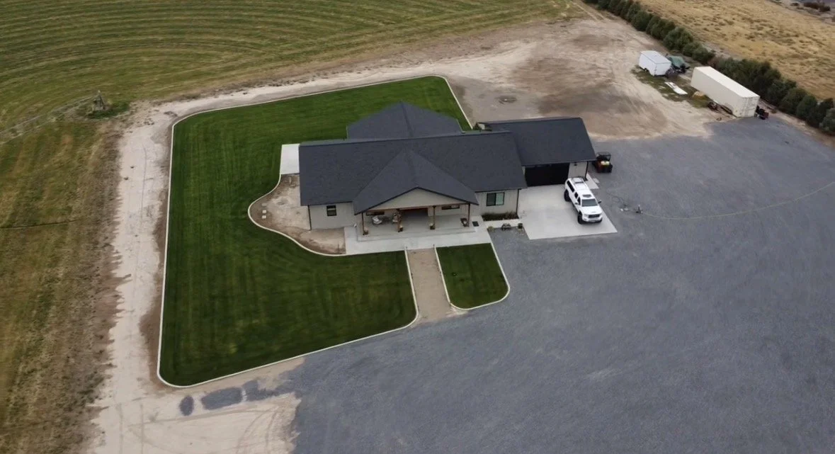 Aerial view of a house with a front porch, surrounded by a large lawn and a gravel driveway, in a rural area.