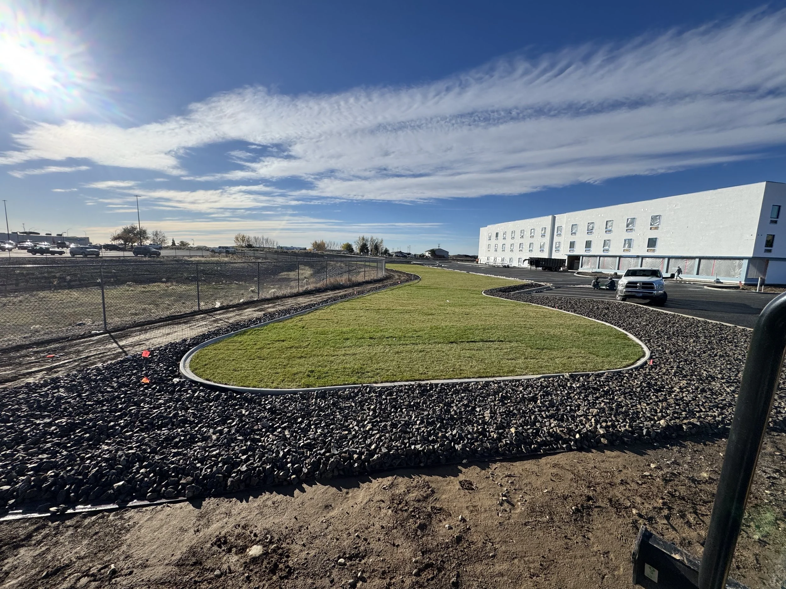 An outdoor scene with a manicured grass area surrounded by black rocks, a chain-link fence, and a large white building with multiple windows in the background. Several cars are parked near the building, and the sky is partly cloudy with the sun shini