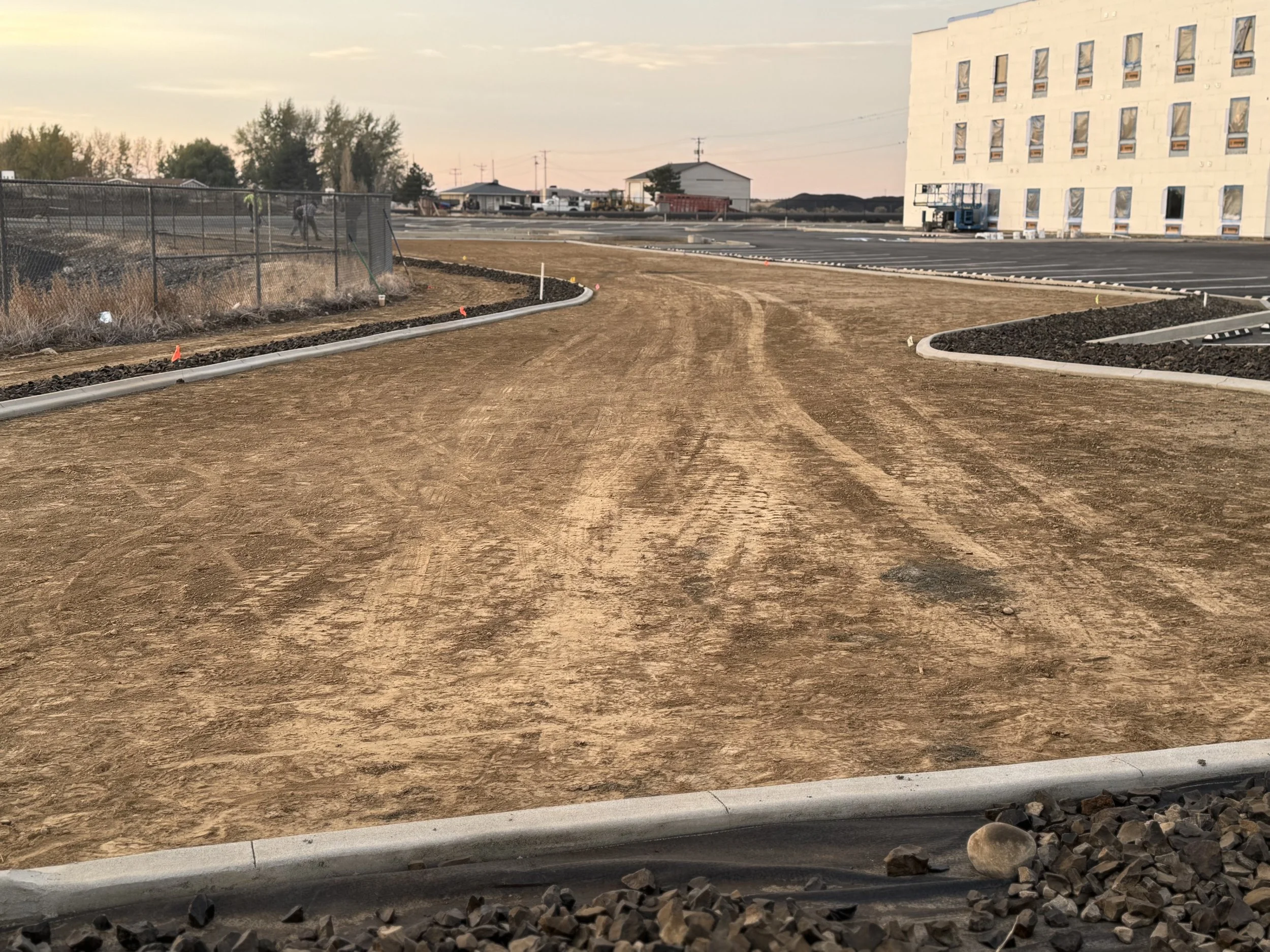 View of a construction site with a dirt road, a fenced area on the left, and a multi-story building on the right, during sunset.