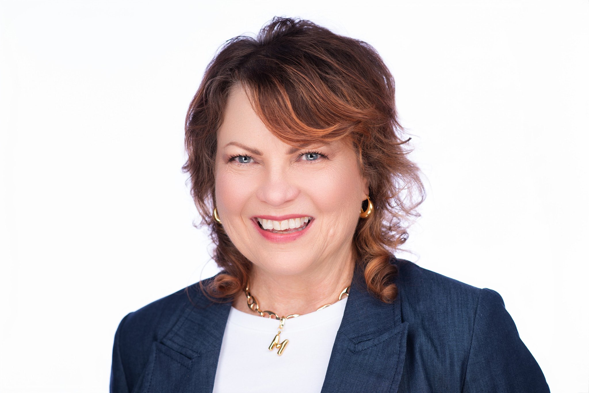 Professional headshot of a woman with shoulder-length auburn hair styled with soft waves, wearing a navy blue blazer over a white top and small pendant necklace. She has a warm, confident smile against a neutral background. Photo by Rojas Photography