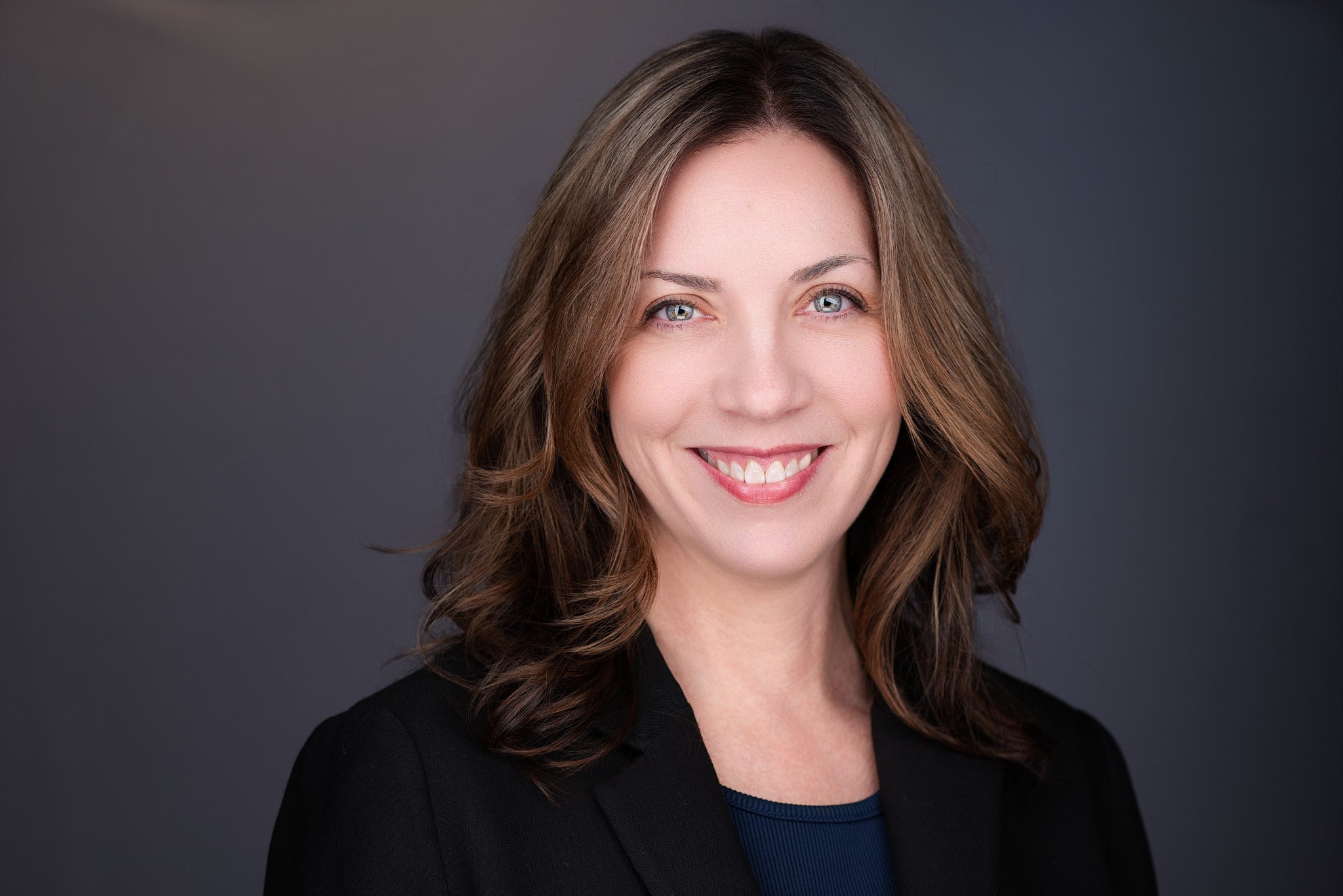 Professional headshot of a smiling woman with shoulder-length brown hair in a business suit on a dark background.