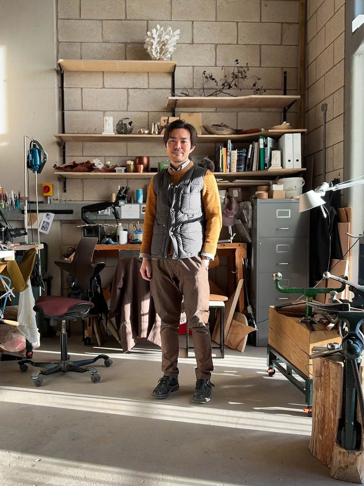Shinta Nakajima standing in a cluttered workshop or studio with shelves, tools, and workbenches.