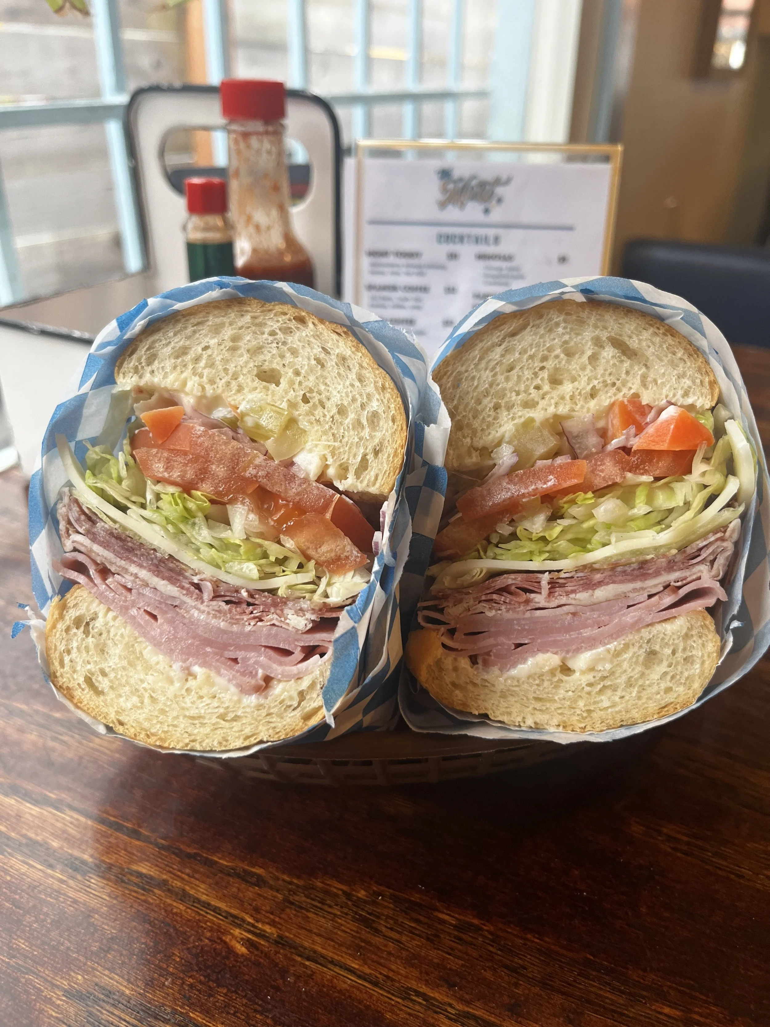 Two sub sandwiches with sliced meats, lettuce, tomato, celery, and mayonnaise in a paper wrapper, on a wooden table in a restaurant. Delicious Sandwiches in Portland Oregon. Lunch