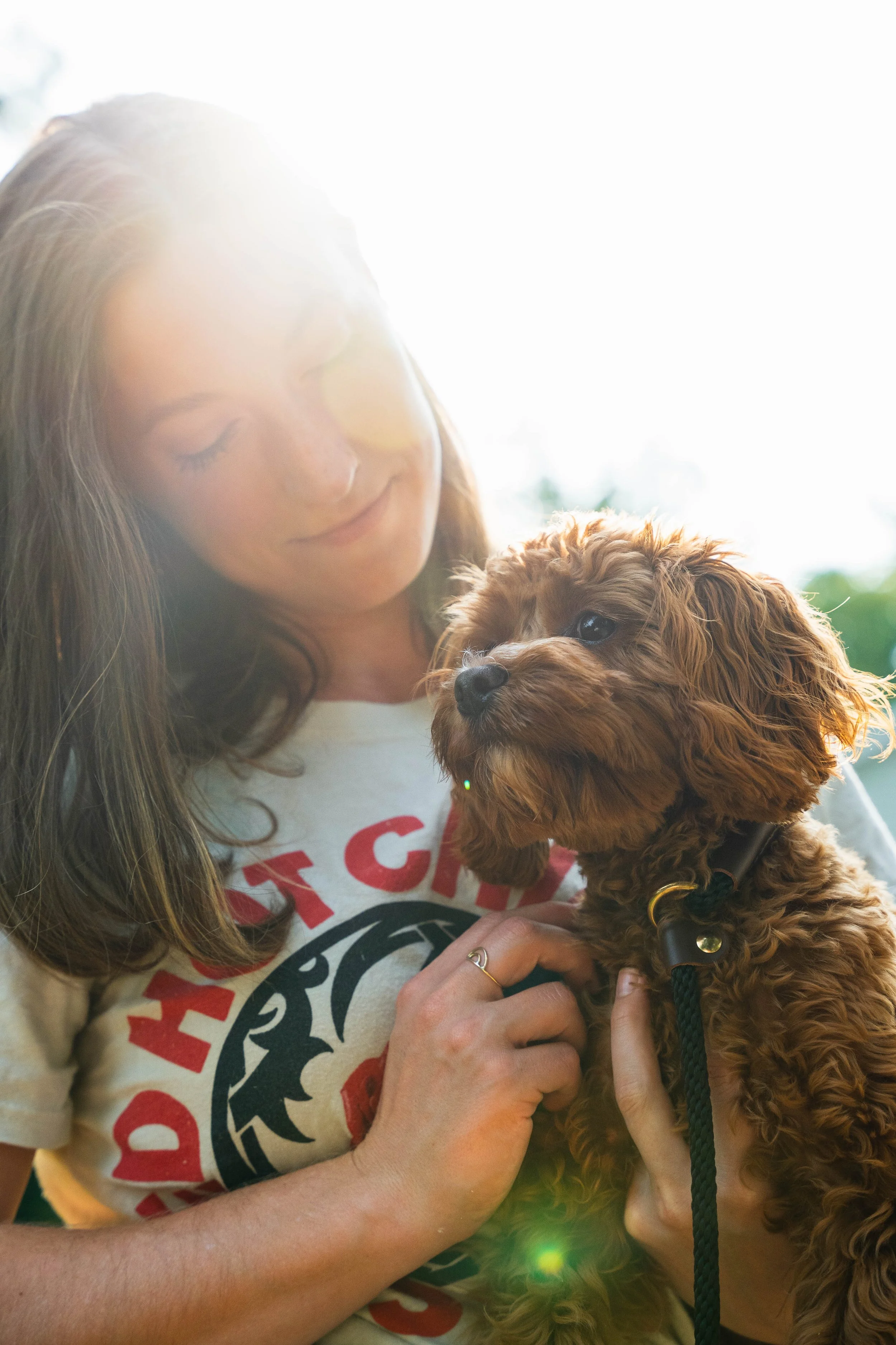 A woman with long brown hair, wearing a beige T-shirt with red and black print, holding a curly-haired small brown puppy with a black collar and leash, both smiling softly.