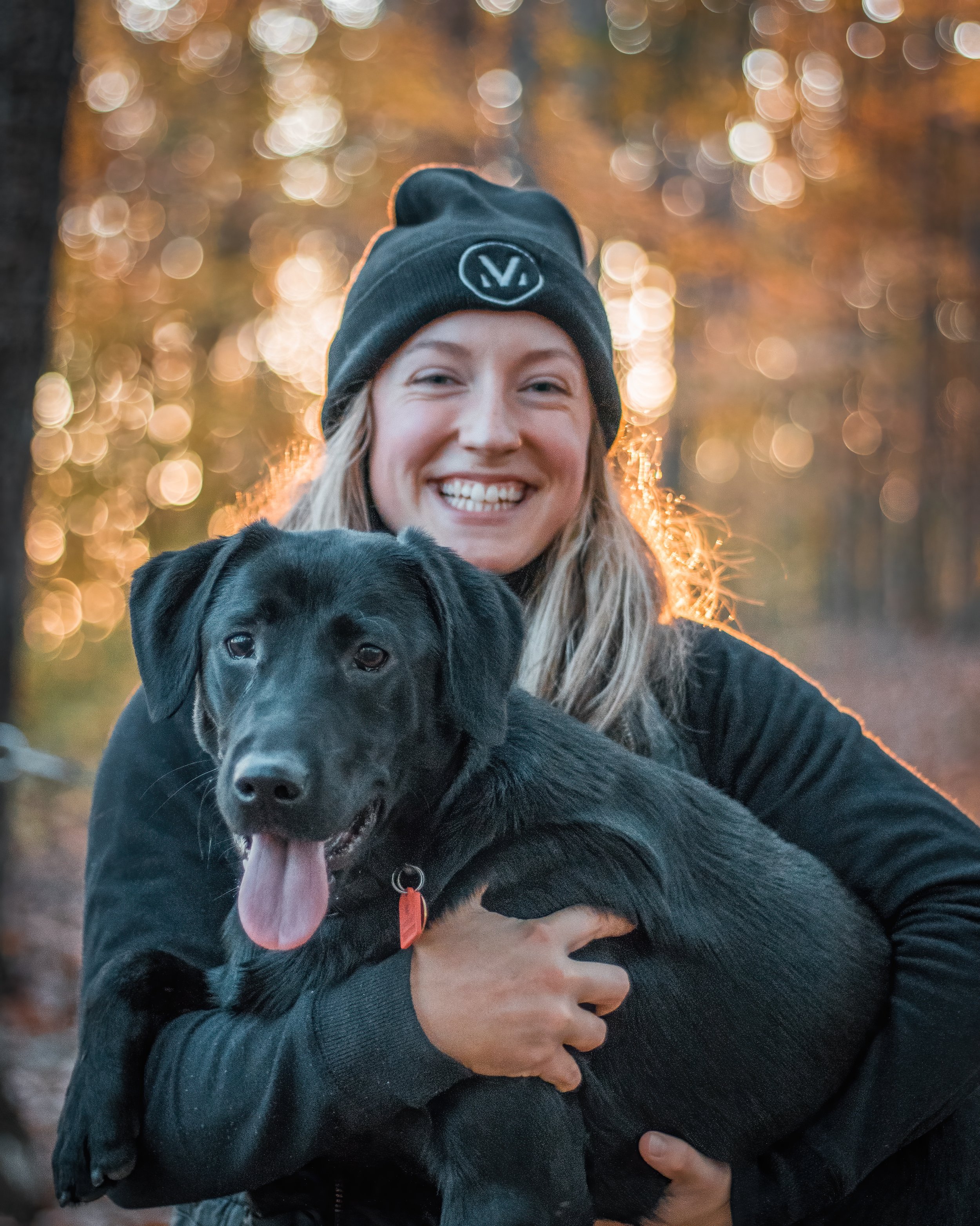 A woman smiling and holding a black puppy outdoors during autumn, with blurred trees and golden leaves in the background.