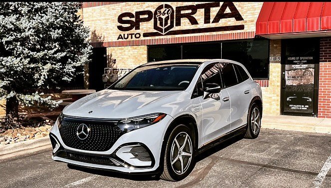 A white Mercedes-Benz EQB SUV parked in front of a storefront with a sign that reads 'SPORTA' and 'AUTO 569' in bold letters, with a red awning and brick building in the background.