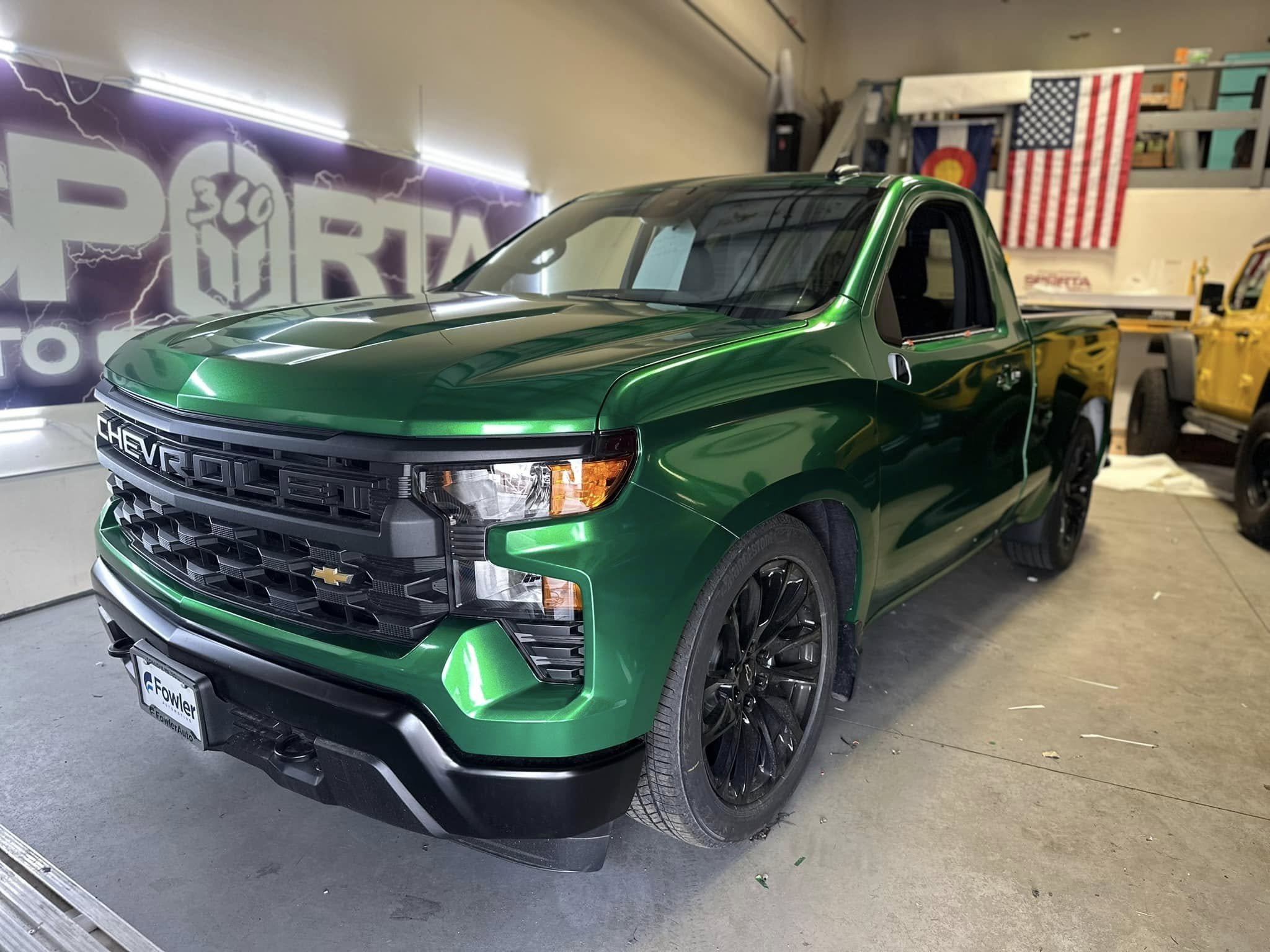 Green Chevrolet pickup truck inside a showroom with American and Colorado flags in the background.