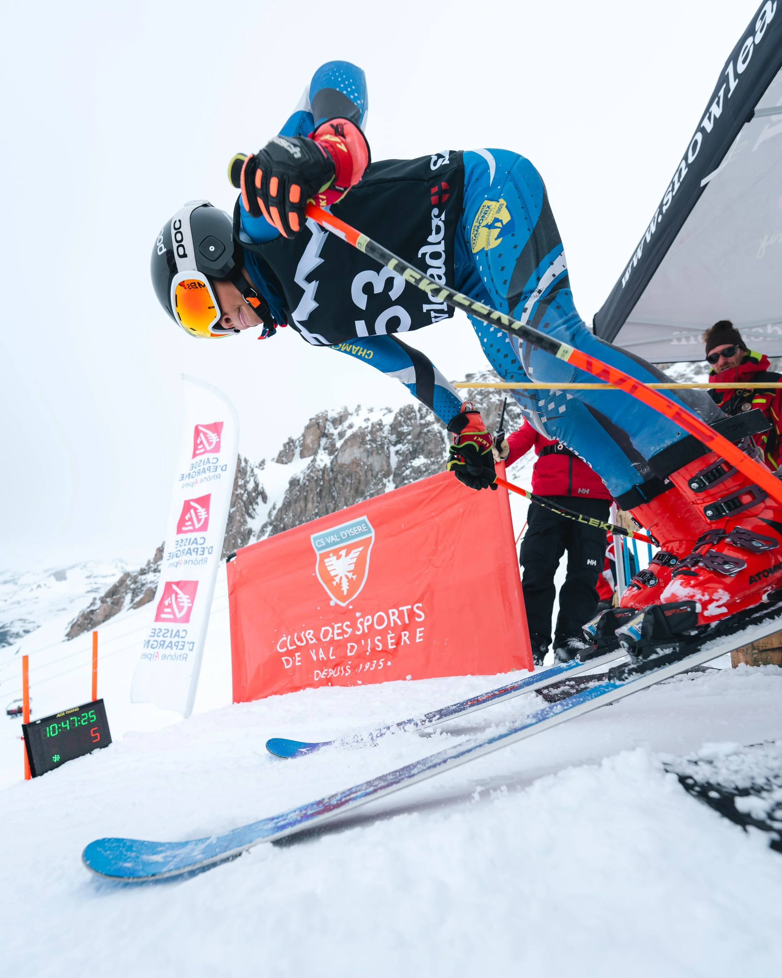 Un skieur au départ d'une course, vêtu d'une combinaison bleue et noire avec un casque noir et des skis rouges, s'élance sur une piste enneigée. En arrière-plan, on aperçoit des bannières de compétition et un panneau du "Club des Sports de Val d'Isèr
