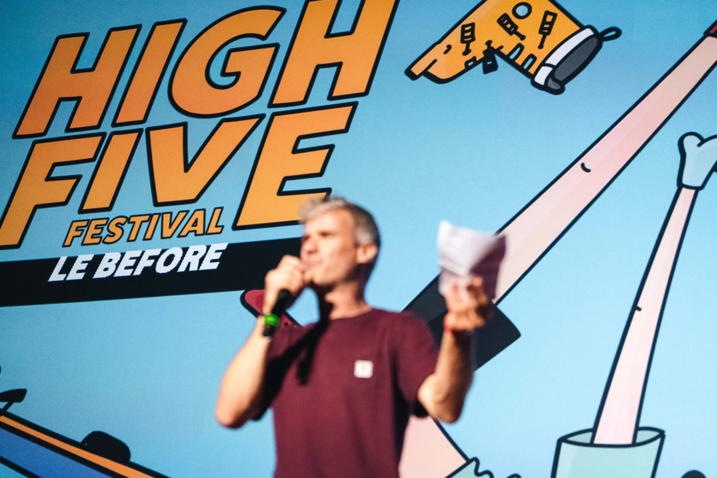 Un homme tenant un micro et des papiers parle devant une affiche du "High Five Festival - Le Before" avec un dessin animé en arrière-plan.