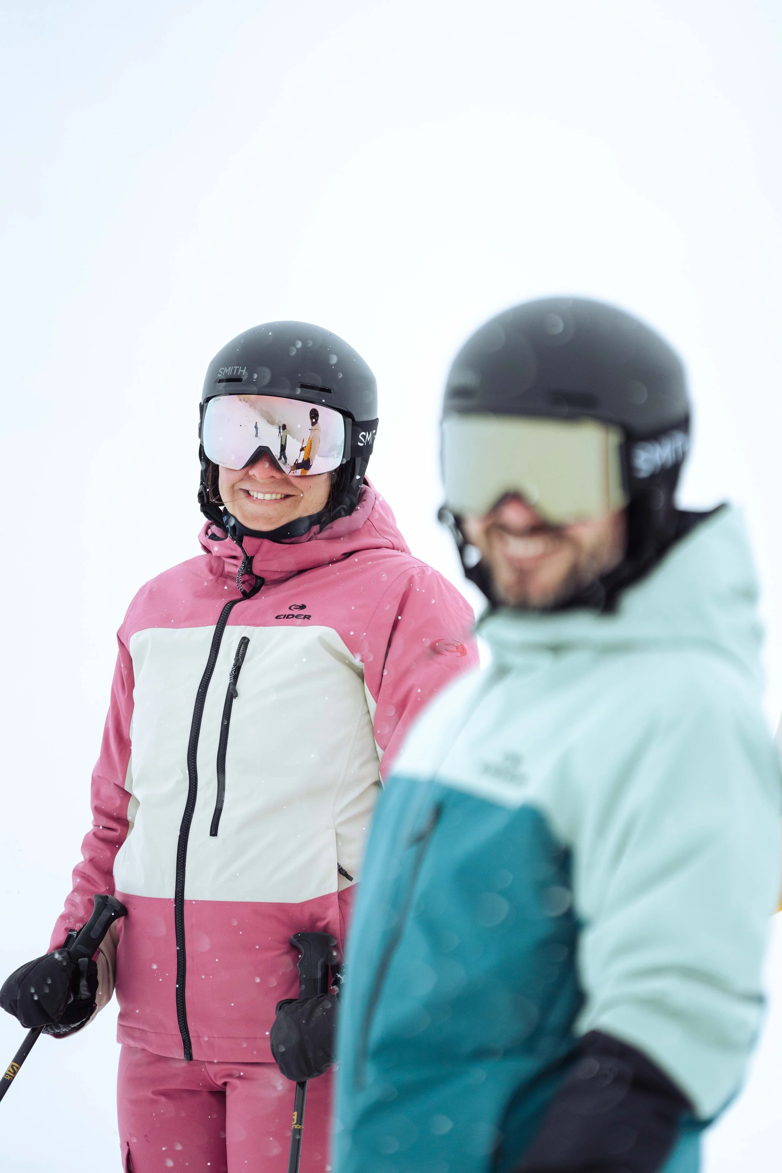 Deux personnes en vêtements de ski avec des casques et des lunettes de protection, tenant des bâtons de ski. Le fond est enneigé.