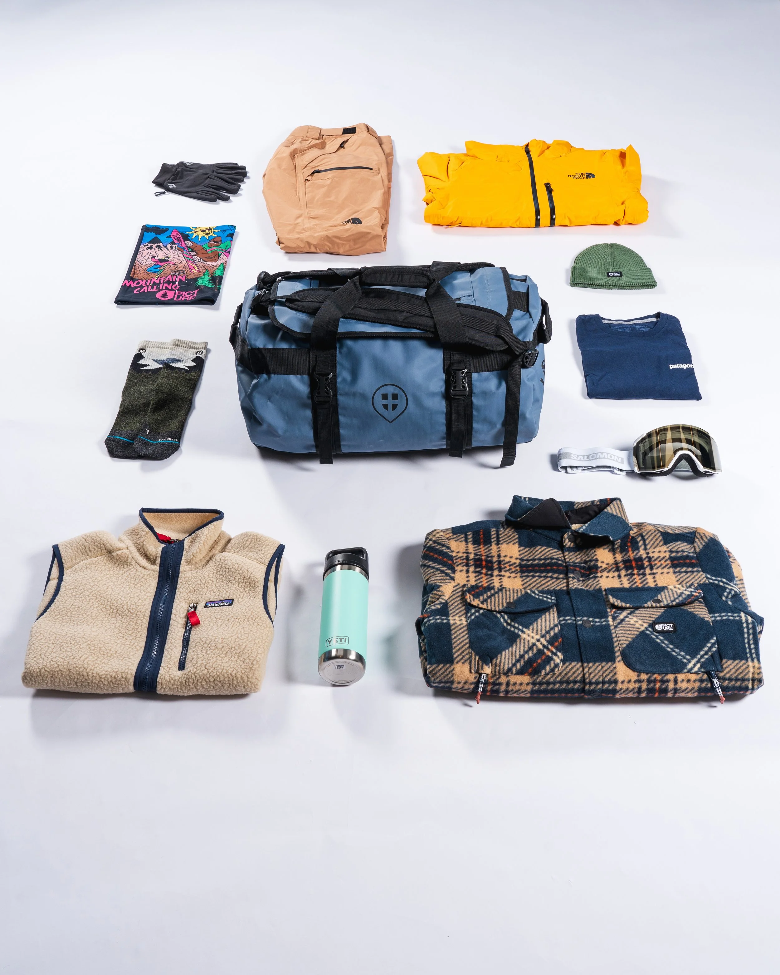 Équipement de plein air comprenant un sac bleu, des vêtements chauds, des gants, des chaussettes, un bonnet vert, un t-shirt bleu, des lunettes de protection, une gourde et des matériaux en laine et polaire, tous disposés sur un fond blanc.