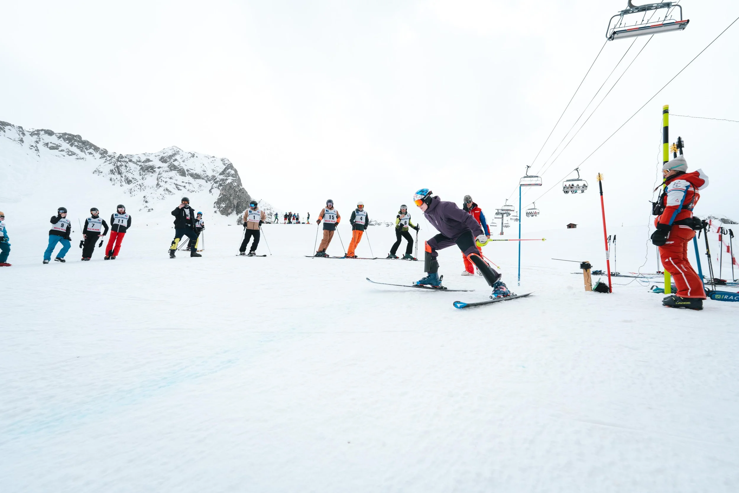 Compétition de ski avec des skieurs prêts à partir, montagnes enneigées et télésiège en arrière-plan.