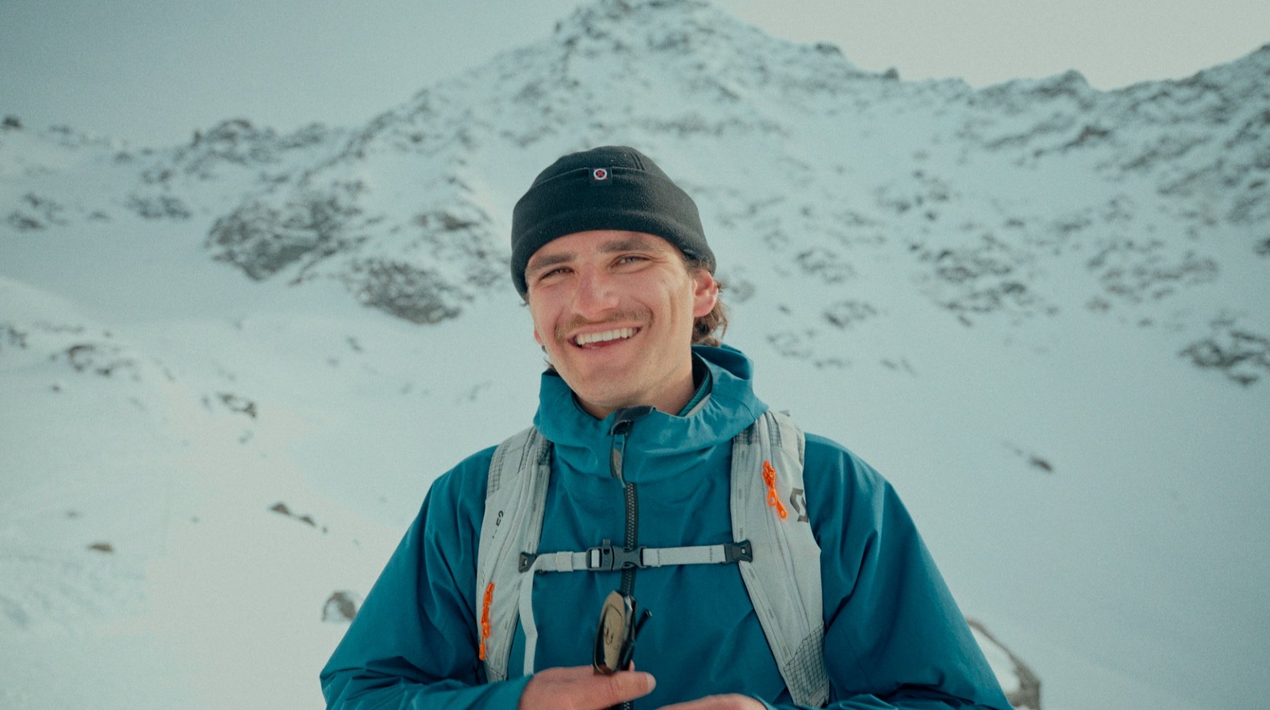 Homme souriant portant une veste bleue et un bonnet noir, debout devant un paysage enneigé de montagne.