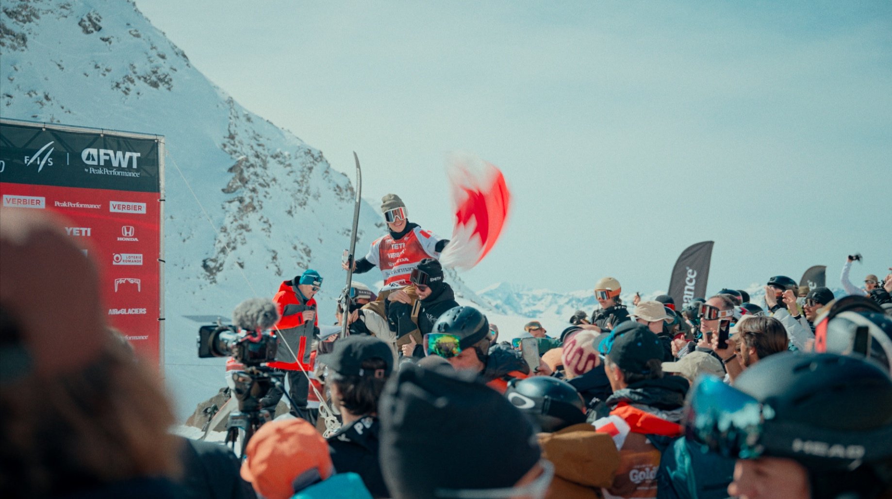 Groupe de personnes avec tenues de ski célébrant en montagne, banderoles et drapeau rouge flou, caméra et équipement de ski visibles, pancarte d'événement avec logos."
