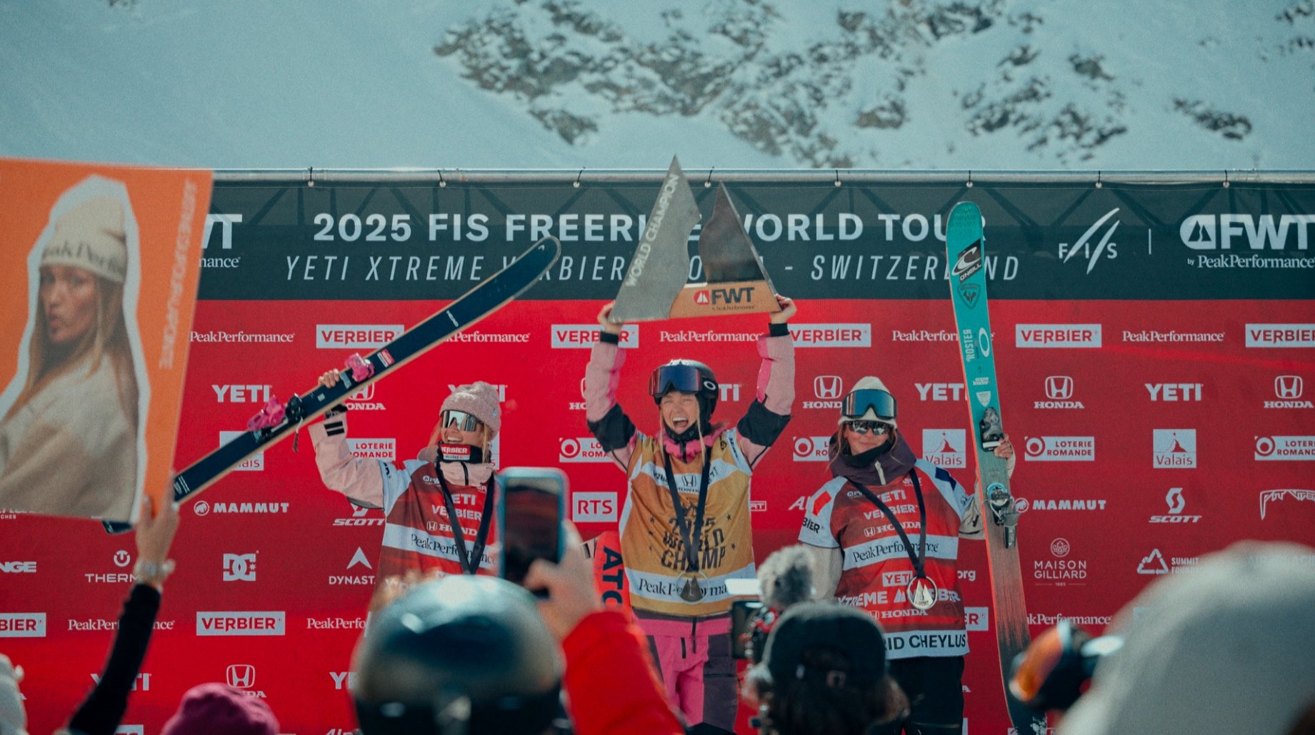 Trois skieuses sur un podium avec des trophées lors du FIS Freeride World Tour 2025 à Verbier, en Suisse, devant un fond de montagnes enneigées.