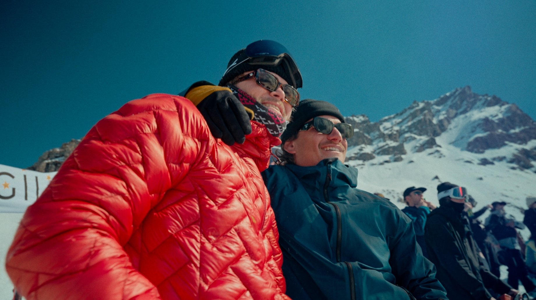 Deux personnes vêtues de vestes de ski sourient sur une montagne enneigée sous un ciel bleu.