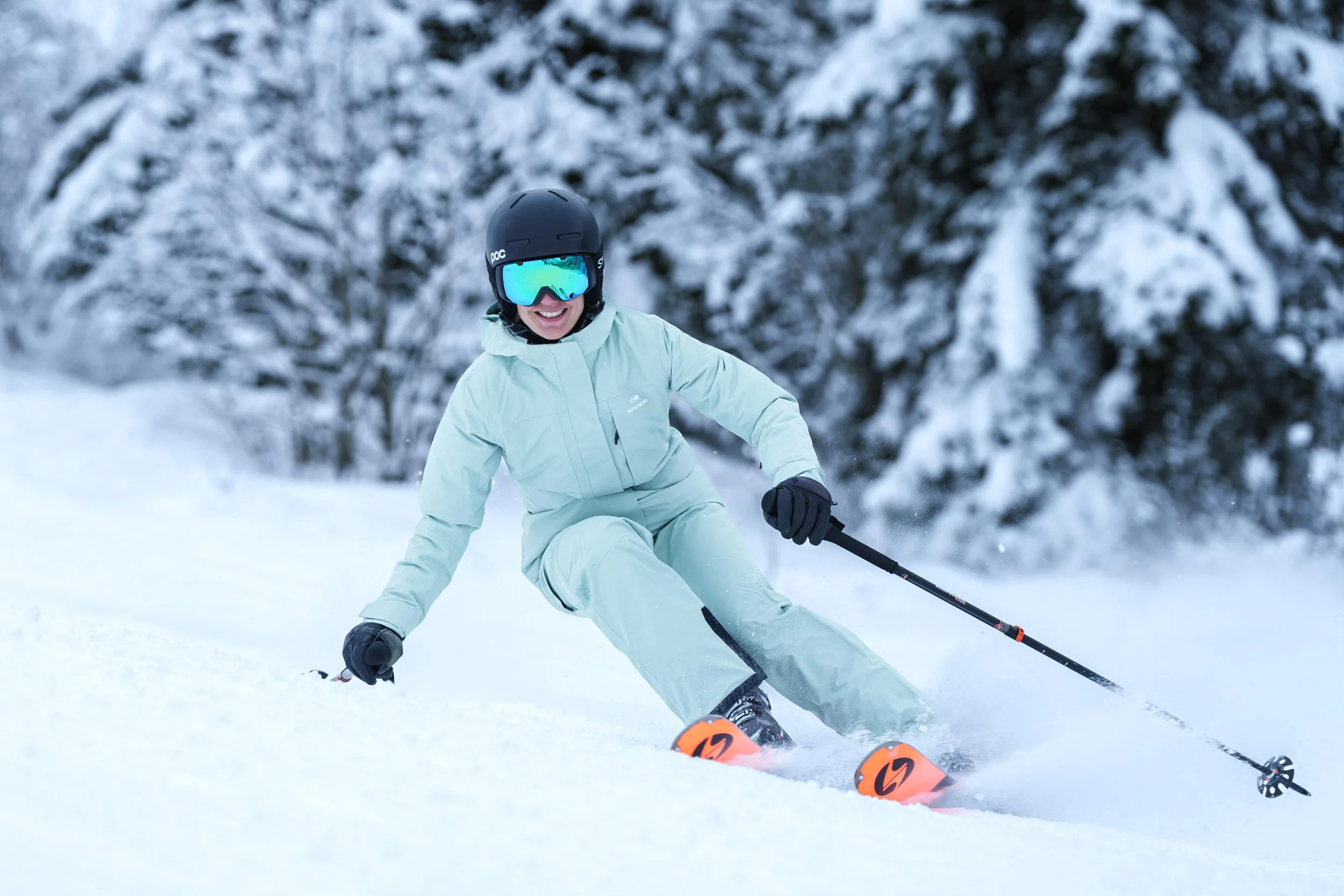 Personne en tenue de ski verte dévalant une pente enneigée avec des skis orange, sous un ciel couvert et des arbres enneigés en arrière-plan.