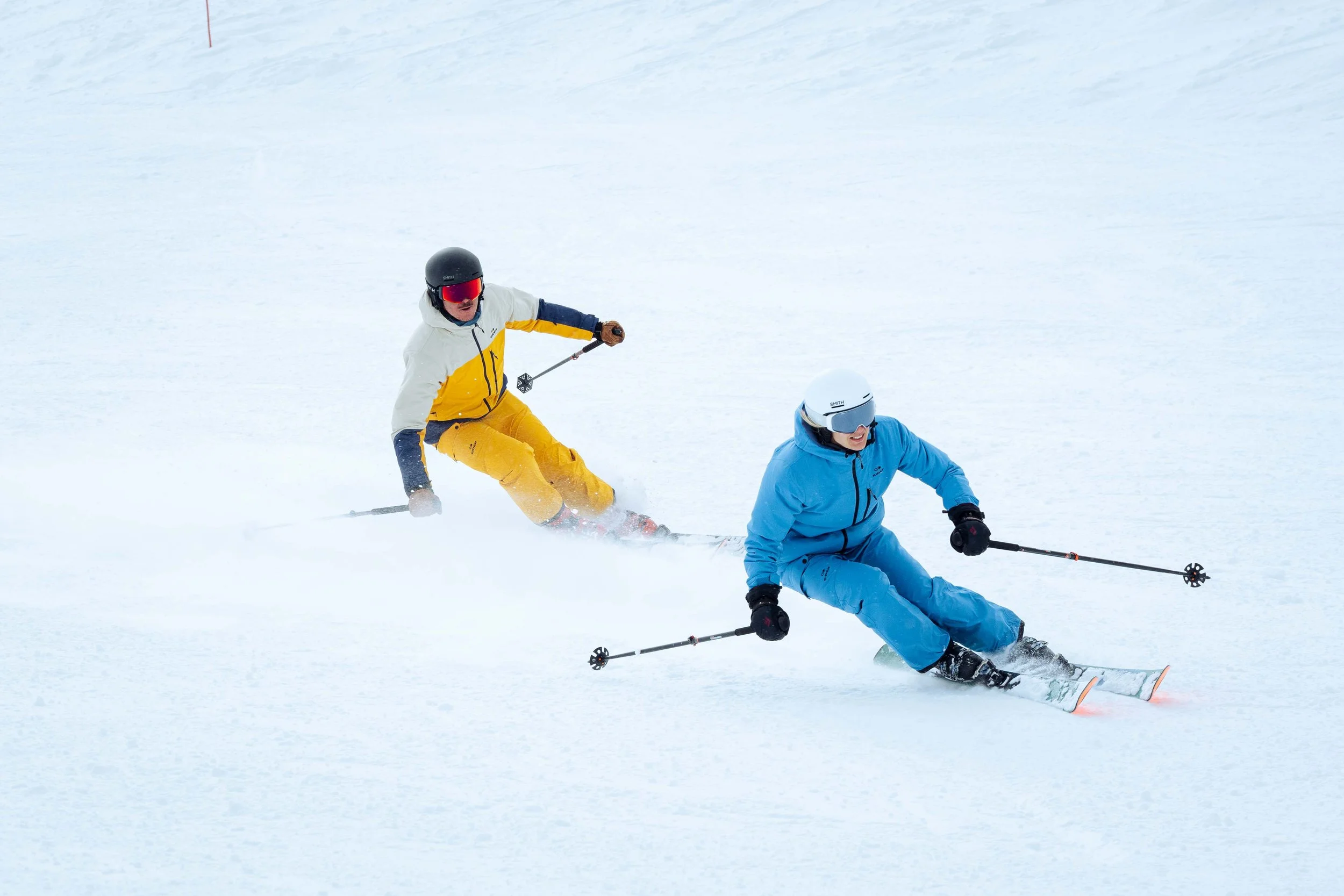 Deux skieurs sur une piste enneigée, portant des combinaisons colorées, l'un en bleu et l'autre en jaune, avec des casques de protection.