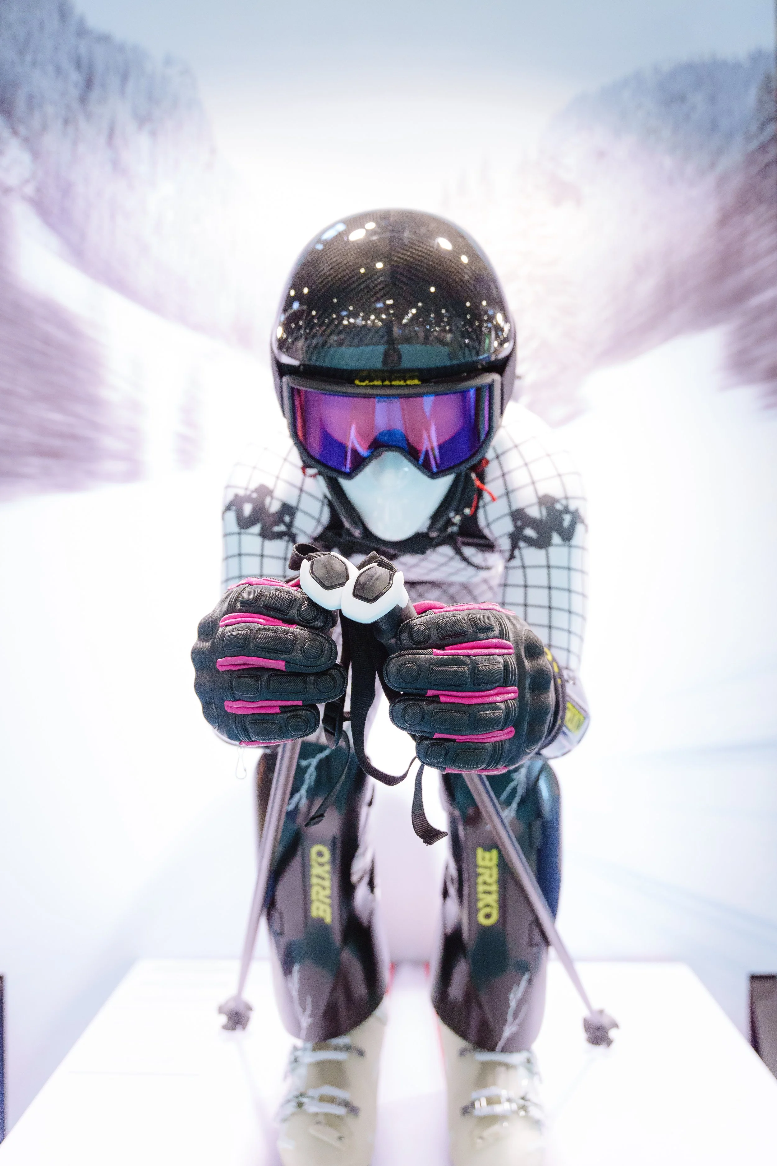 Un mannequin de ski habillé en tenue de ski aérodynamique, portant un casque et des lunettes, tenant des bâtons de ski, devant un fond enneigé flou.