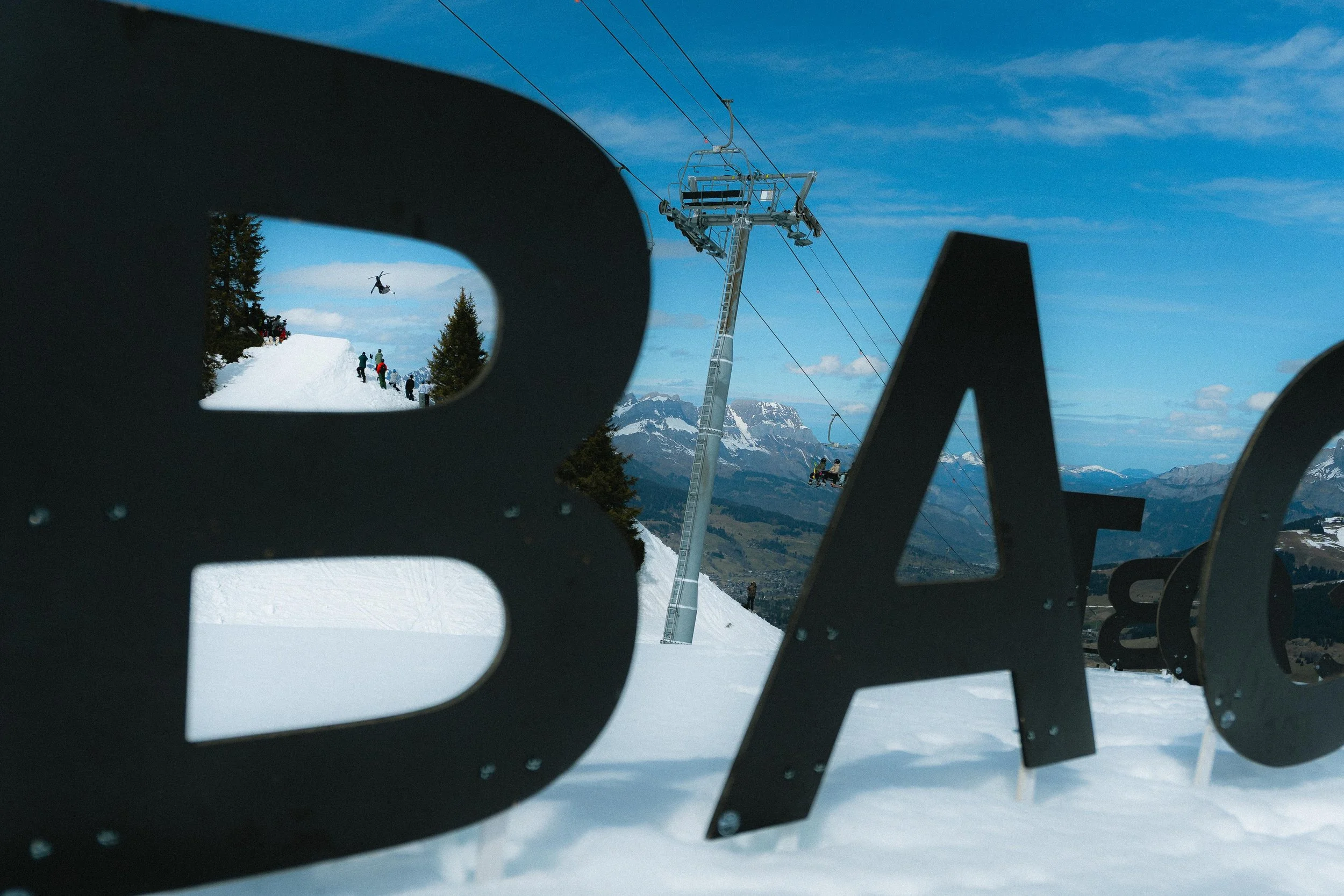 Paysage de montagne enneigée avec un skieur en action, un télésiège, et des lettres en métal au premier plan, sous un ciel bleu.