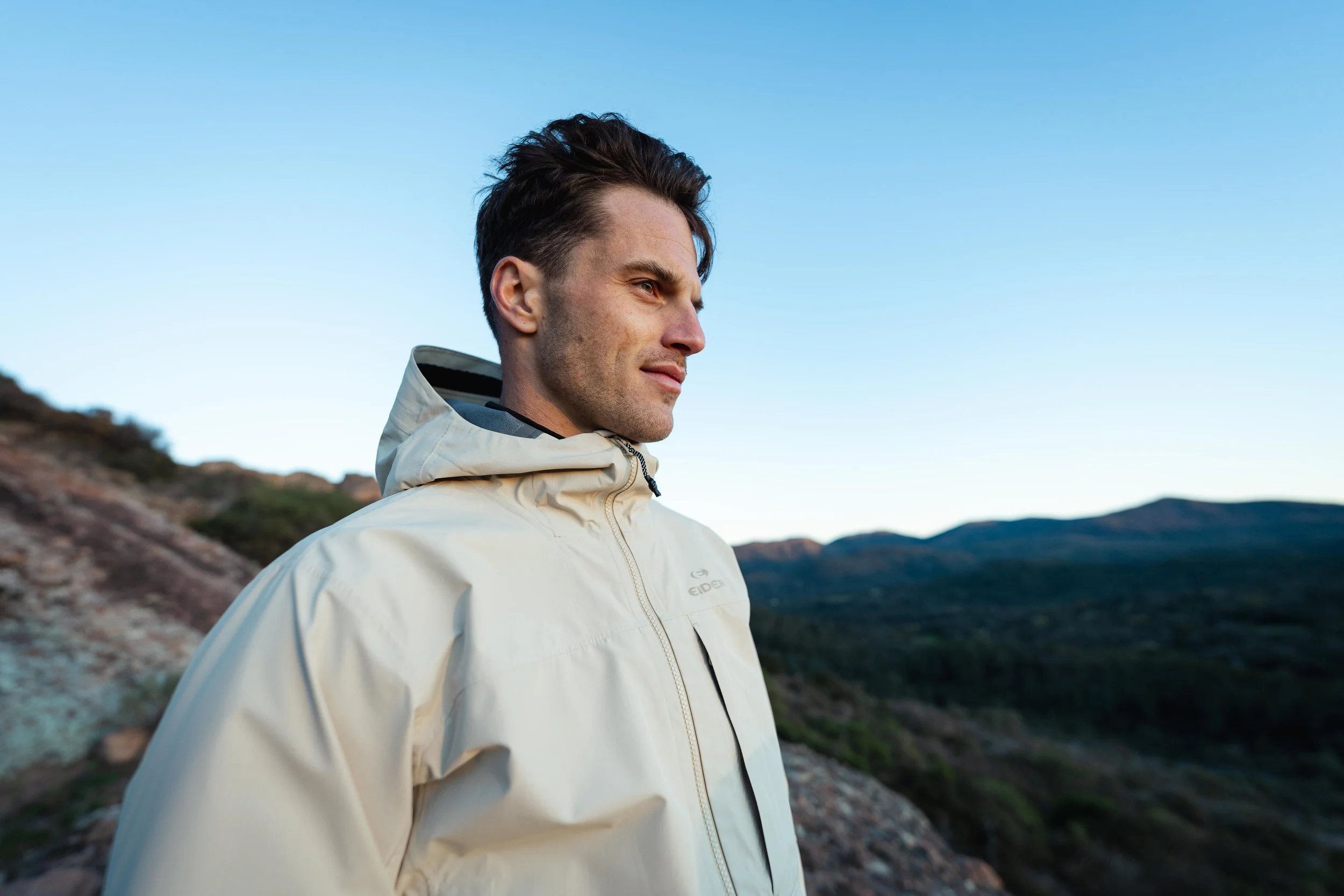 Un homme en veste beige regarde au loin dans un paysage de montagnes sous un ciel bleu clair.