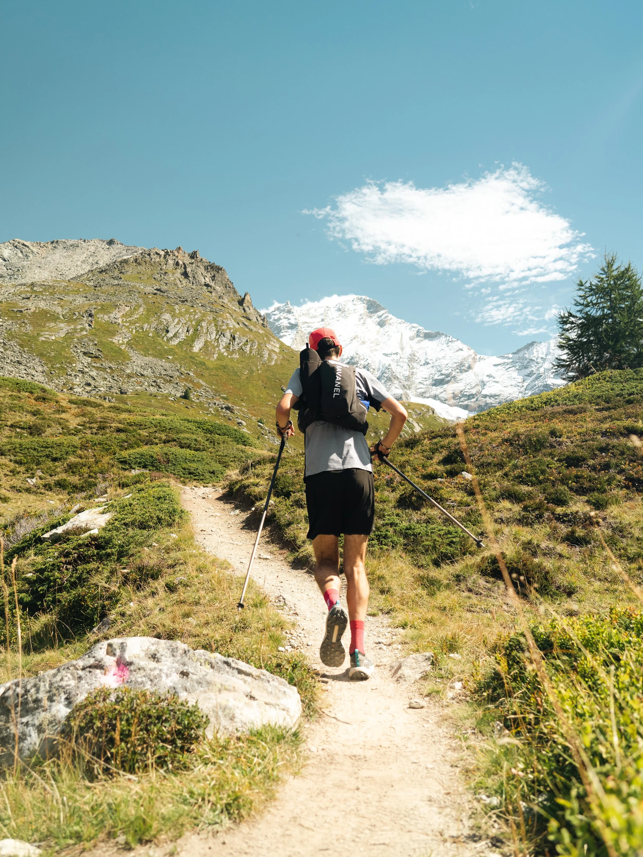 Une personne fait de la randonnée en montagne avec des bâtons de marche, portant un sac à dos et une casquette rouge, sur un sentier entouré de végétation. En arrière-plan, des montagnes enneigées sont visibles sous un ciel bleu avec quelques nuages.
