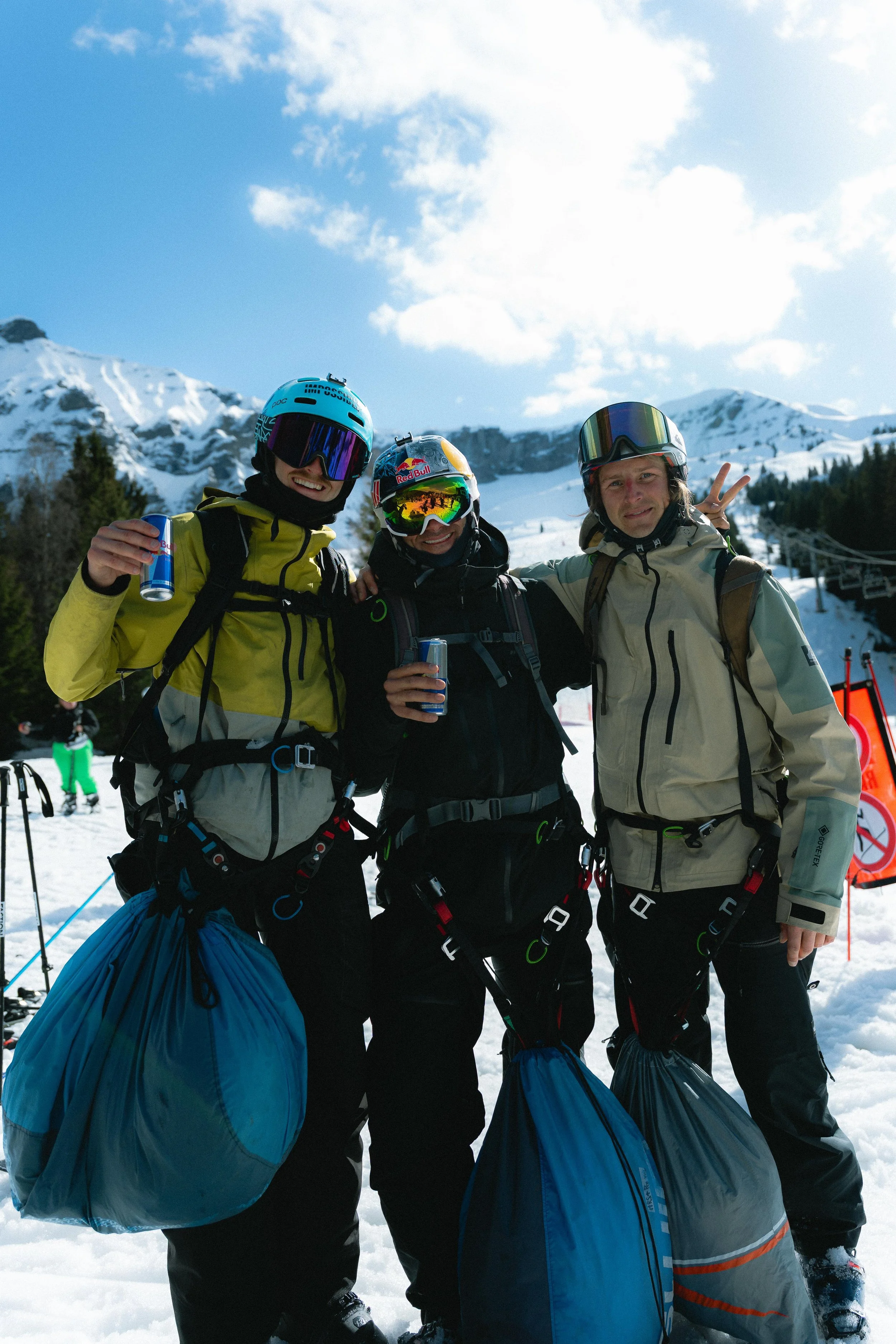 Trois skieurs souriants portant des casques et lunettes de ski, tenant des boissons énergétiques, avec des parapentes sur neige et montagnes en arrière-plan.