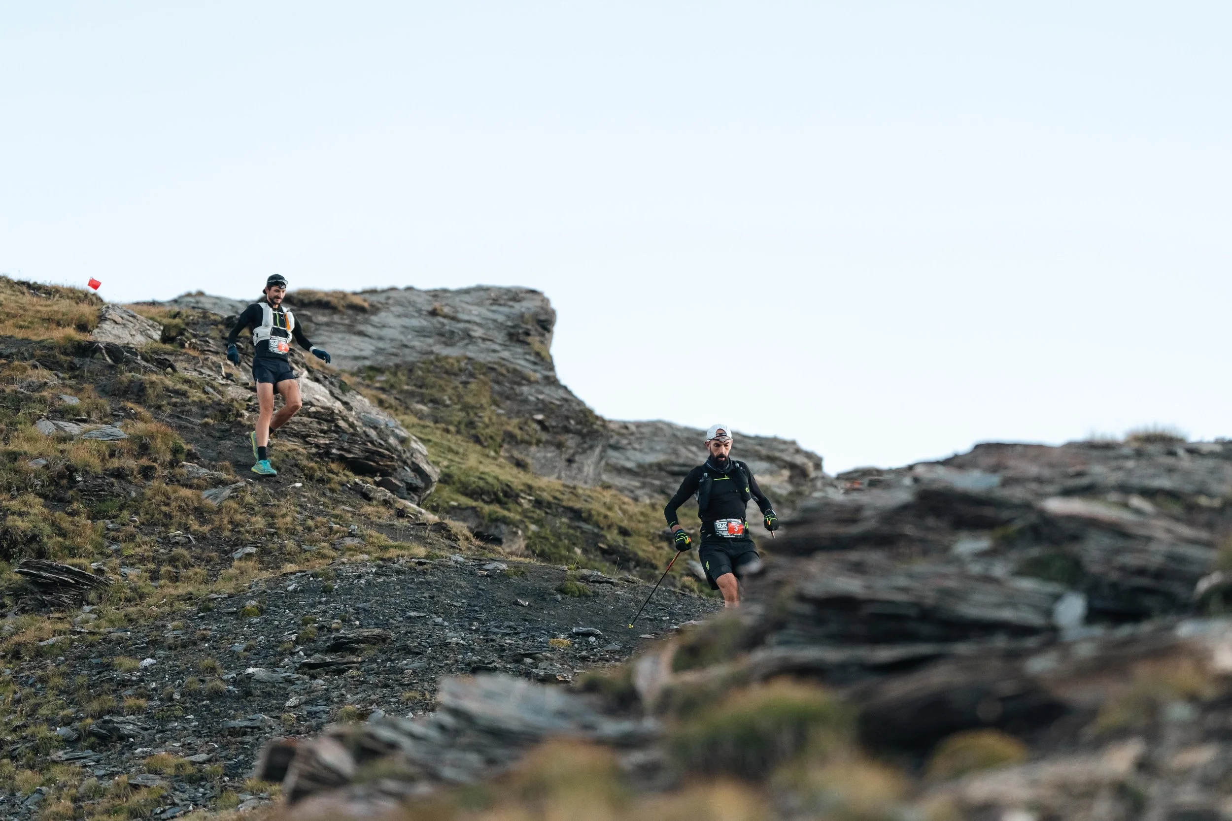 Deux coureurs descendent un sentier rocailleux en montagne, portant des vêtements de sport et des équipements de trail.