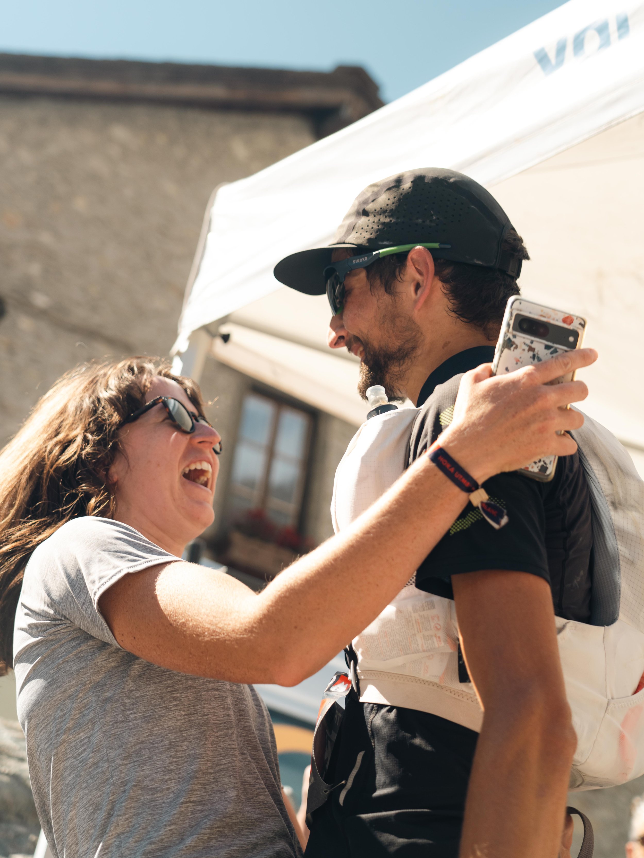 Deux personnes portant des lunettes de soleil rient et partagent un moment joyeux en extérieur. L'une tient un téléphone et l'autre porte un sac à dos léger. Il y a une tente et un bâtiment en arrière-plan.