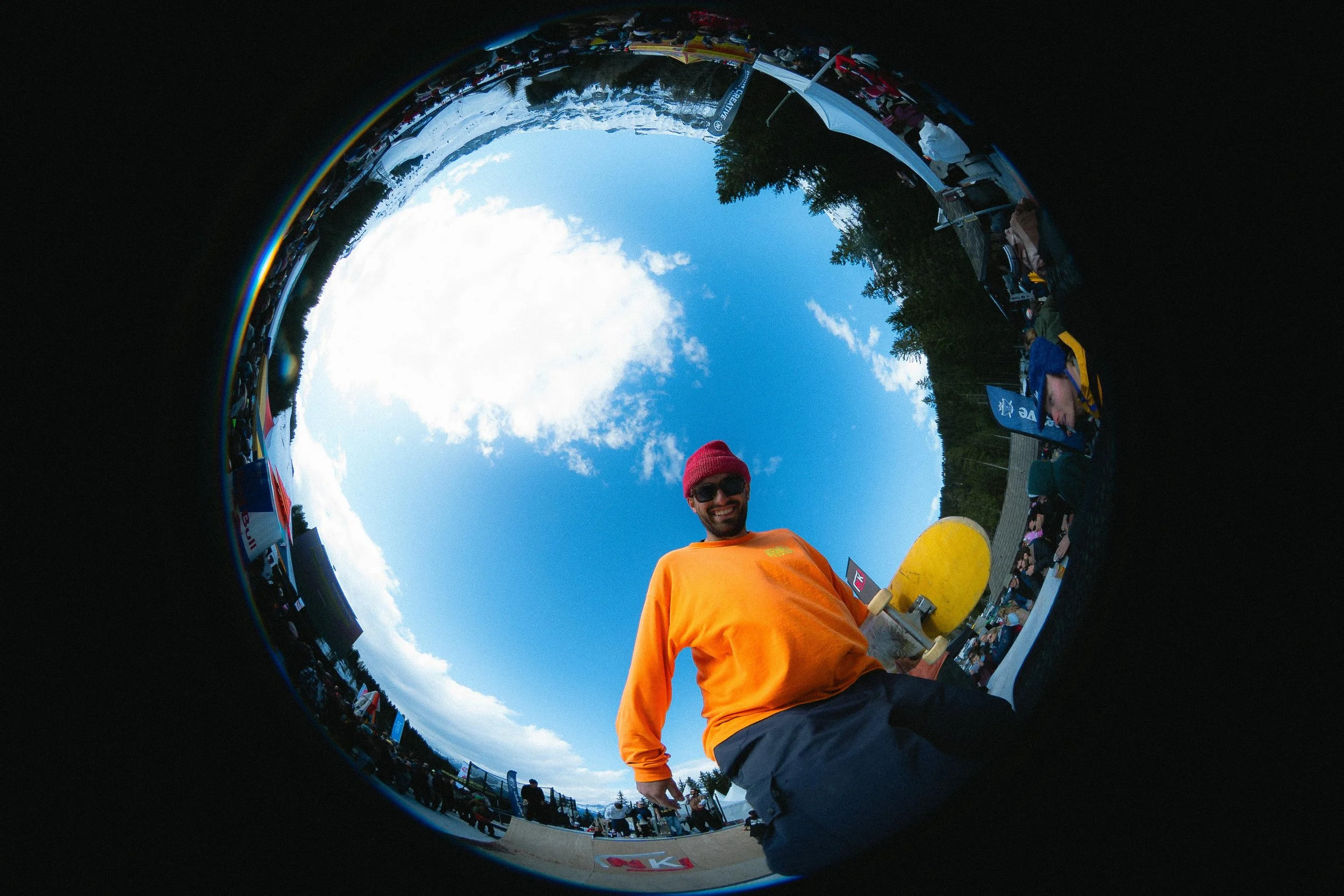 Homme en t-shirt orange tenant un skateboard, photographié à travers un objectif fish-eye, ciel bleu et rassemblement en arrière-plan.