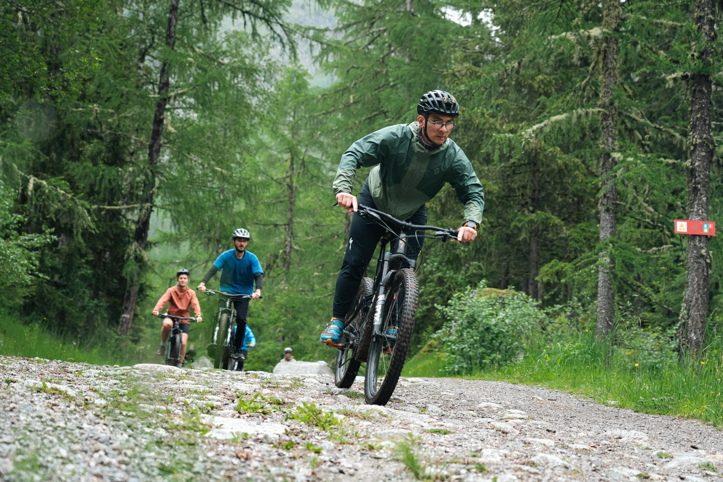 Quatre cyclistes portant des casques roulant sur un chemin de terre dans une forêt verdoyante.