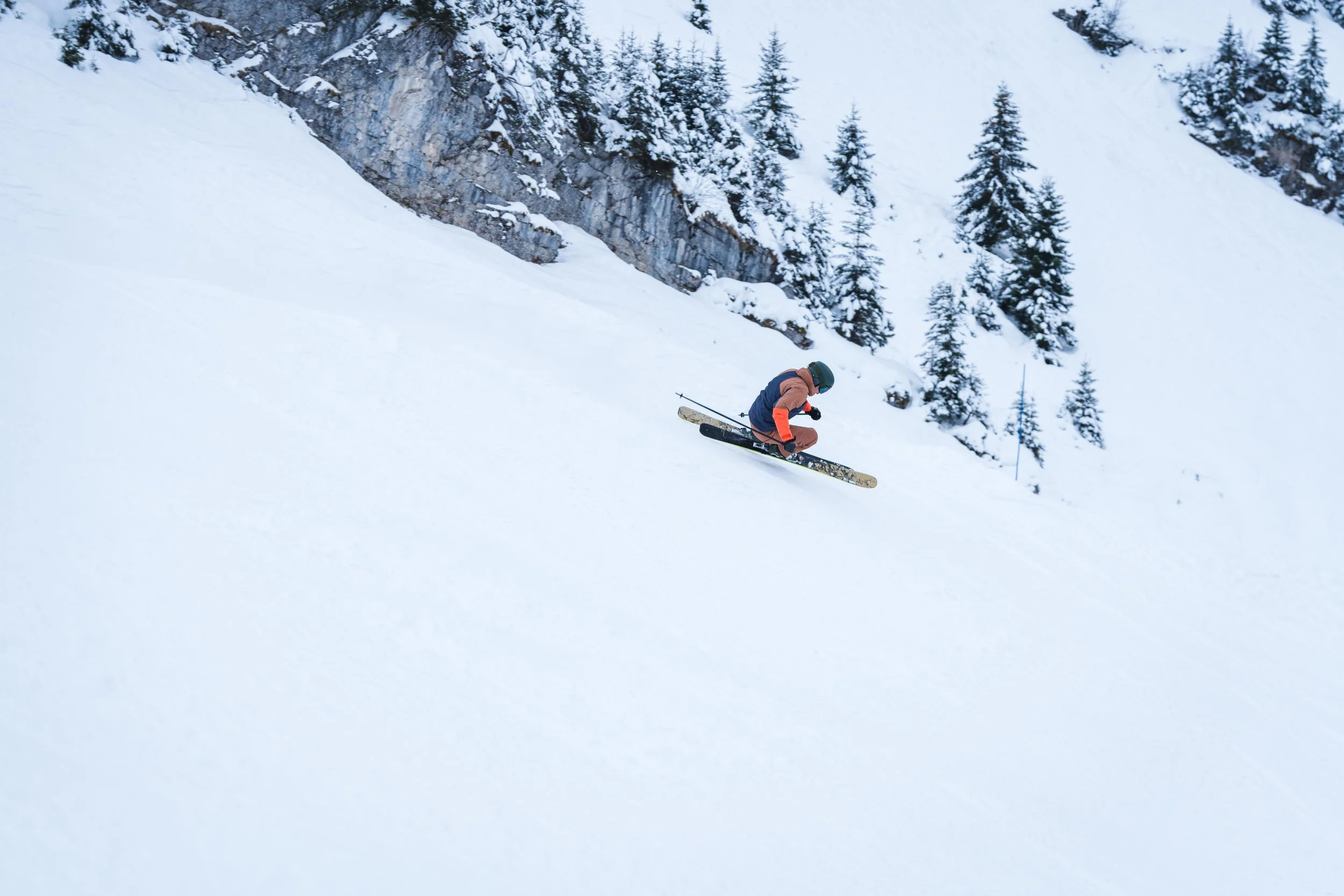 Une personne faisant du ski sur une pente enneigée, entourée de sapins et de falaises.