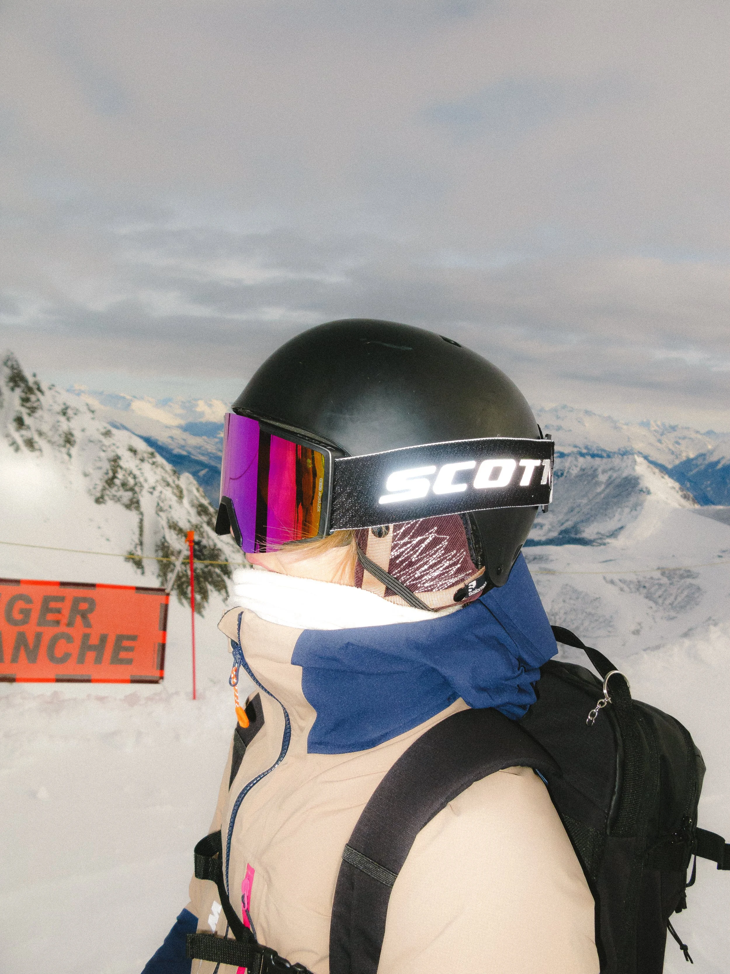 Personne portant un casque de ski noir avec des lunettes de protection colorées, vêtue d'une veste beige et bleu, avec un sac à dos, se tient sur une piste enneigée. Un panneau "Danger Avalanche" se trouve à côté. Les montagnes sont visibles en arriè