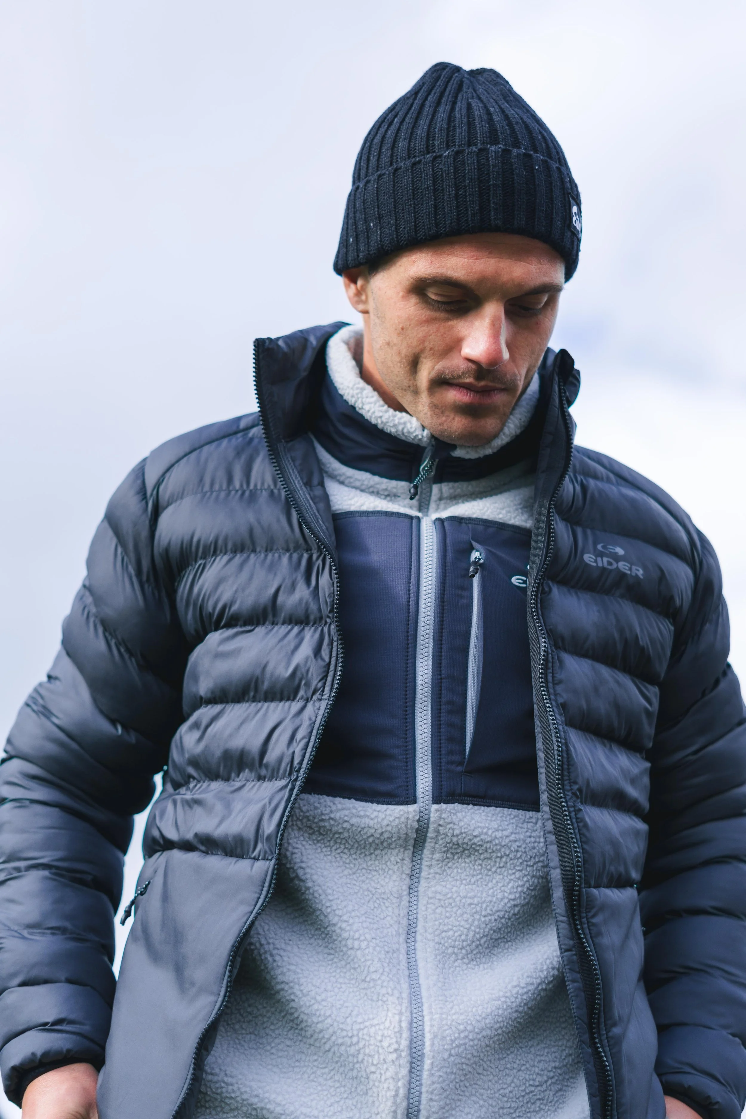 Un homme portant une doudoune noire, une veste polaire grise, et un bonnet noir.