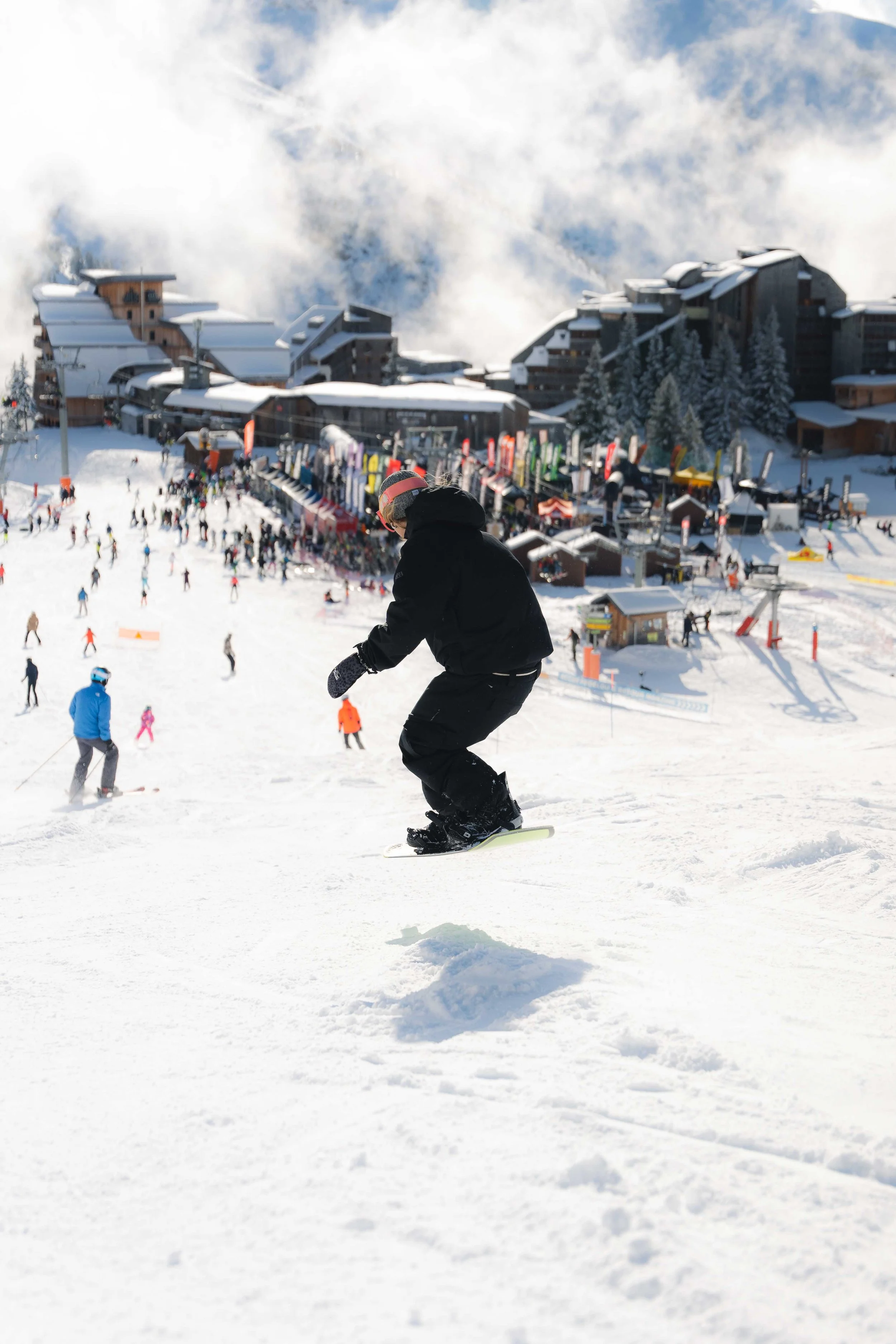 Personne faisant du snowboard sur une piste neigeuse avec des bâtiments et d'autres skieurs en arrière-plan.
