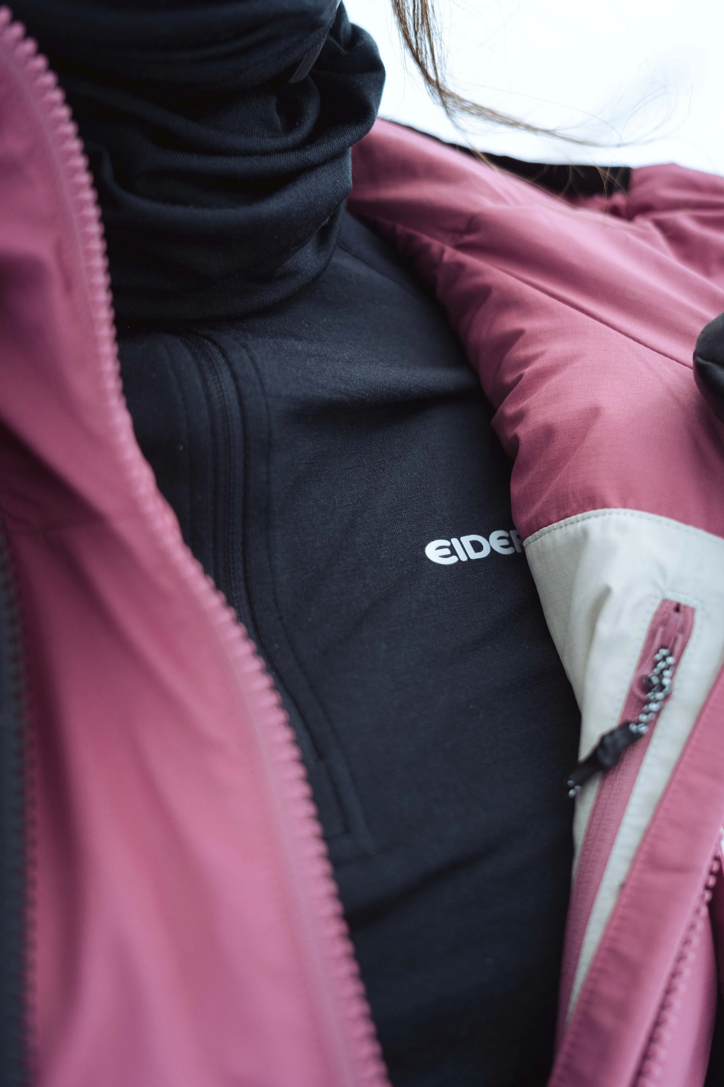 Personne portant un manteau rose avec une veste noire sous. Marques visibles incluant "Eider" sur le vêtement noir.